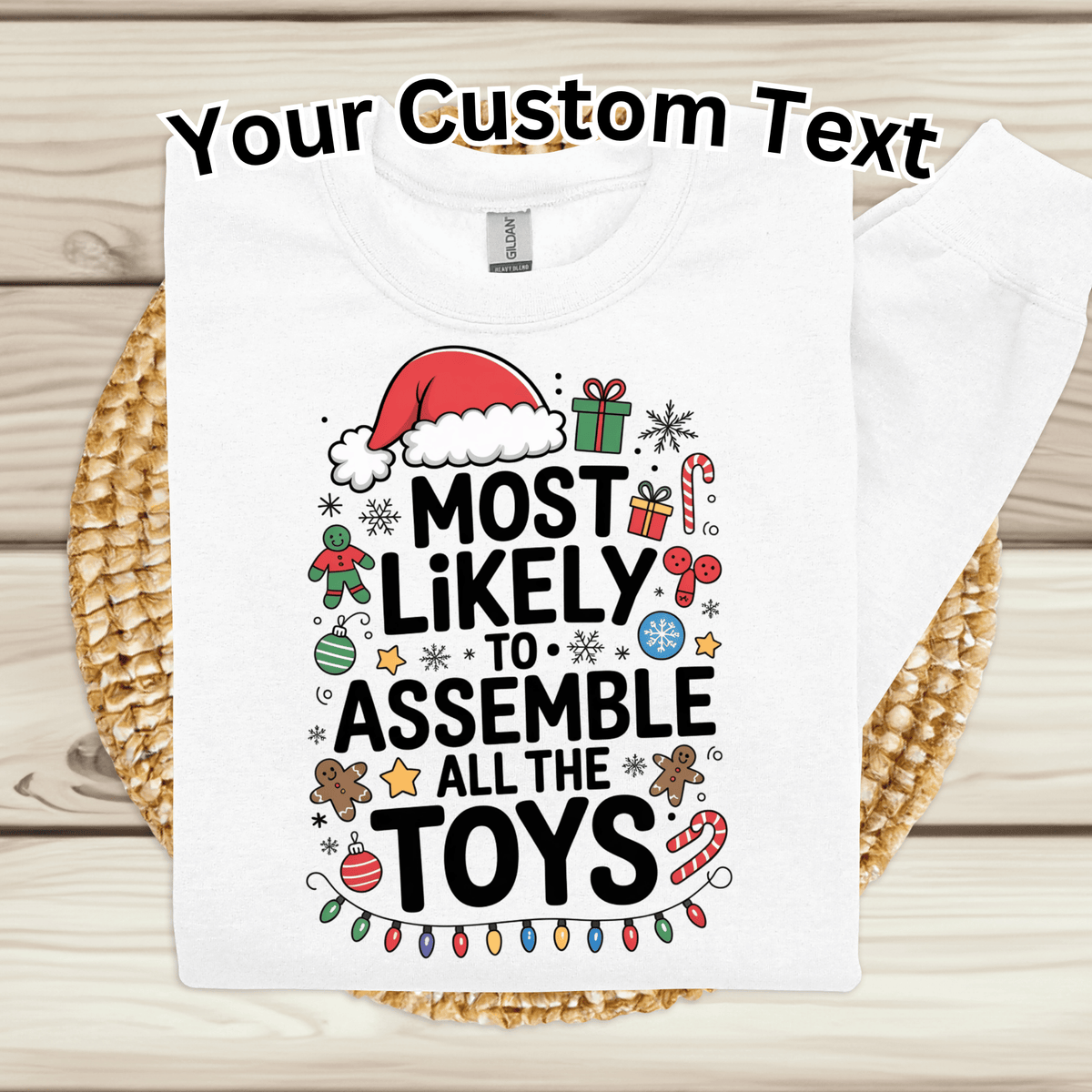 A folded white T-shirt laid flat on a woven placemat over a light wood surface. The shirt has a large black slogan reading 'Most Likely to Assemble All the Toys' surrounded by colorful Christmas-themed icons including a Santa hat, candy canes, ornaments, and gingerbread cookies.