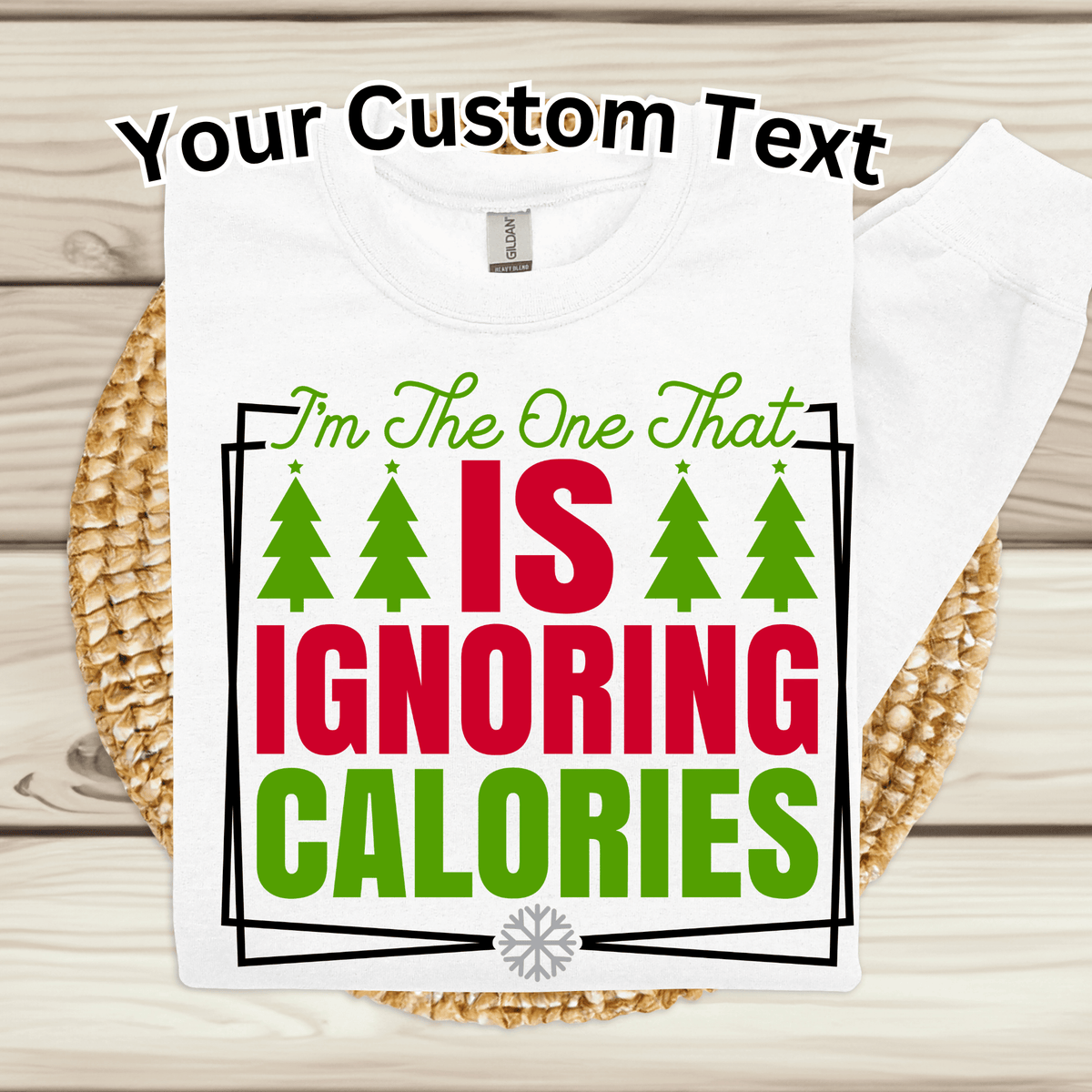 A white custom graphic t-shirt displayed on a woven placemat over a light wood background. The shirt features a festive graphic with the text 'I'm The One That IS IGNORING CALORIES' in green and red, decorated with four green Christmas trees.