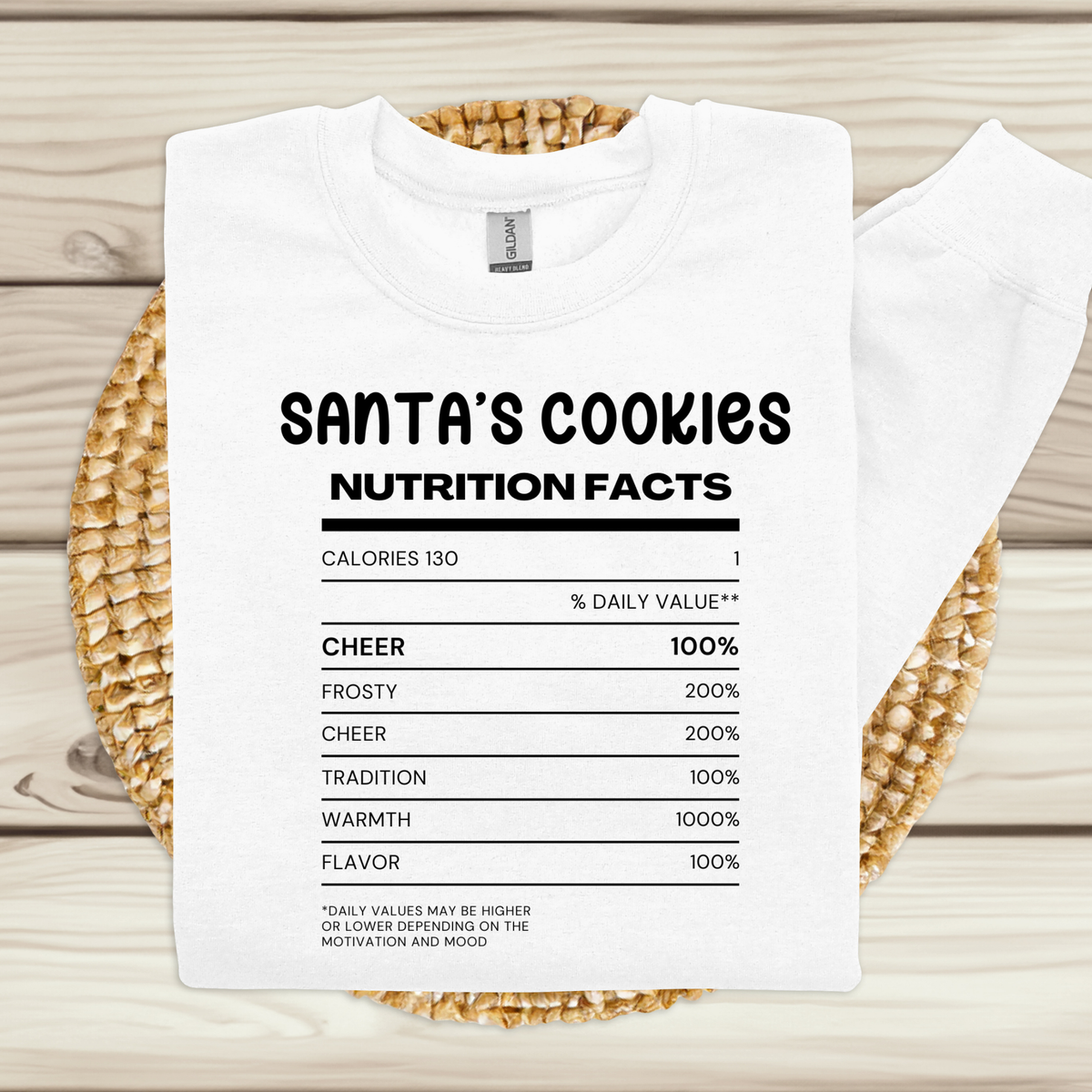 A white crewneck sweatshirt with a black printed 'Santa's Cookies' nutrition facts graphic, laid flat on a woven straw mat over a light wood background.