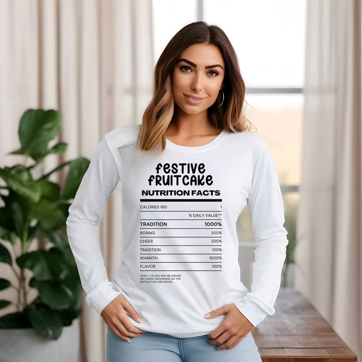 A woman wearing a white long-sleeve t-shirt with a black graphic print that reads 'festive fruitcake nutrition facts' above a fake nutrition label, posed in a bright indoor setting with curtains and a plant.