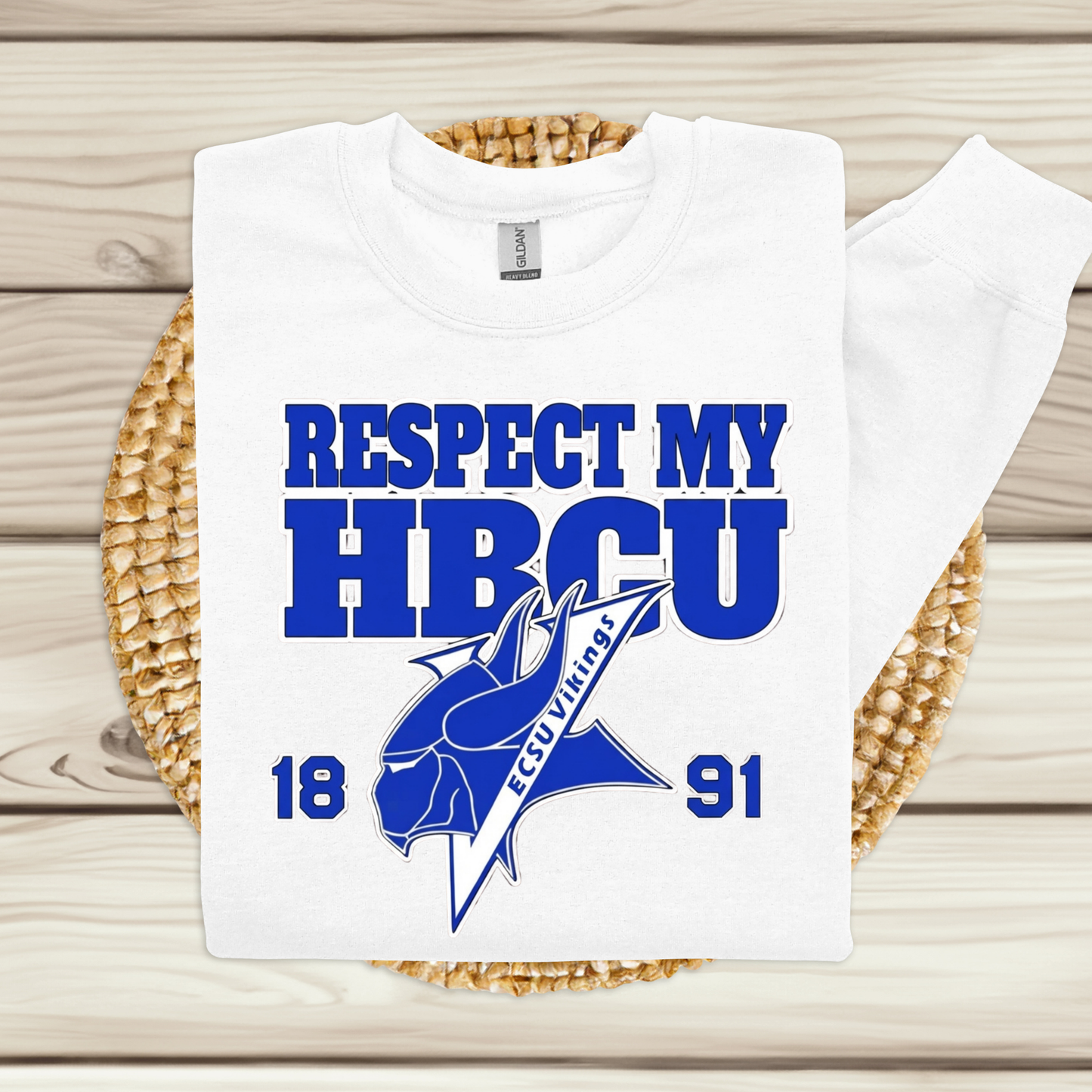 A white long-sleeve T-shirt laid flat on a woven round mat over a light wood plank surface. The shirt has a large blue graphic reading 'RESPECT MY HBCU' with a fish illustration and 'ECU VALUABLE' text, plus the numbers 18 and 91.