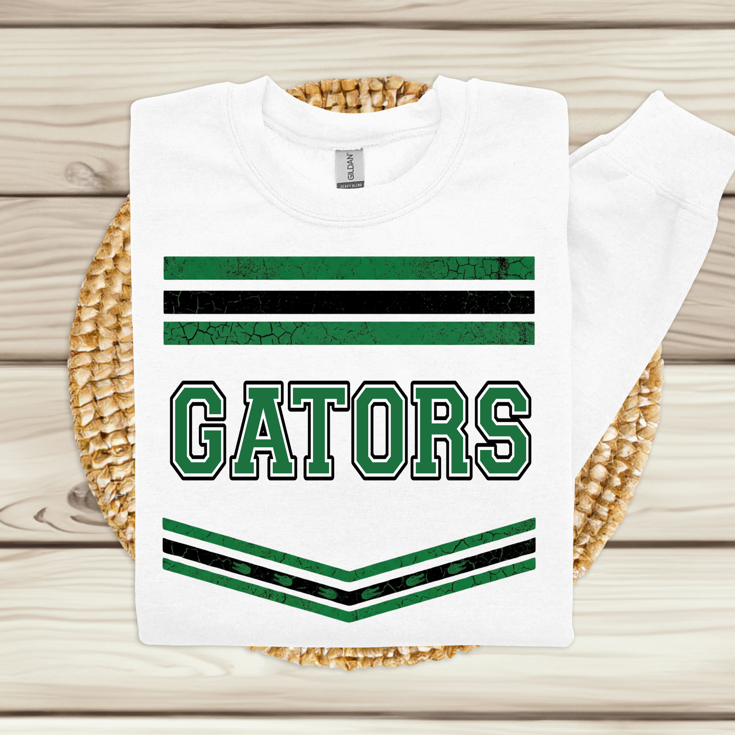 A white long-sleeve sweatshirt with green and black stripes and the word 'GATORS' printed on the front, displayed on a woven round mat over a light wood plank background.