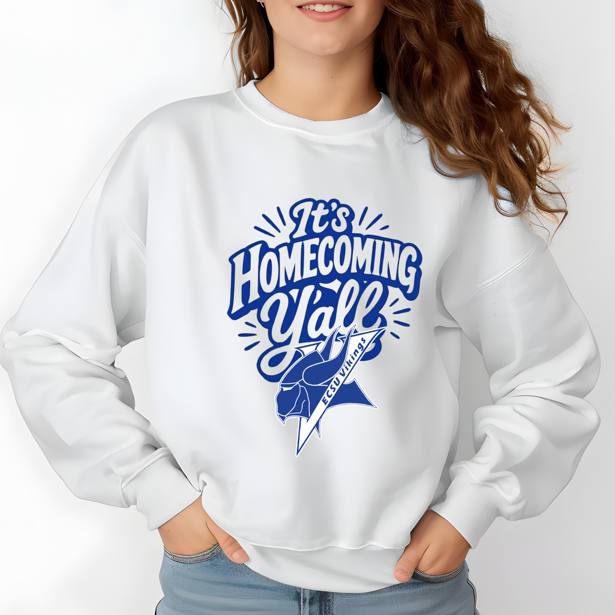 A woman wearing a white crewneck sweatshirt with a blue graphic that reads 'It's Homecoming Y'all' and 'BY THE NUMBERS' with a ribbon graphic, paired with light blue jeans against a white background.