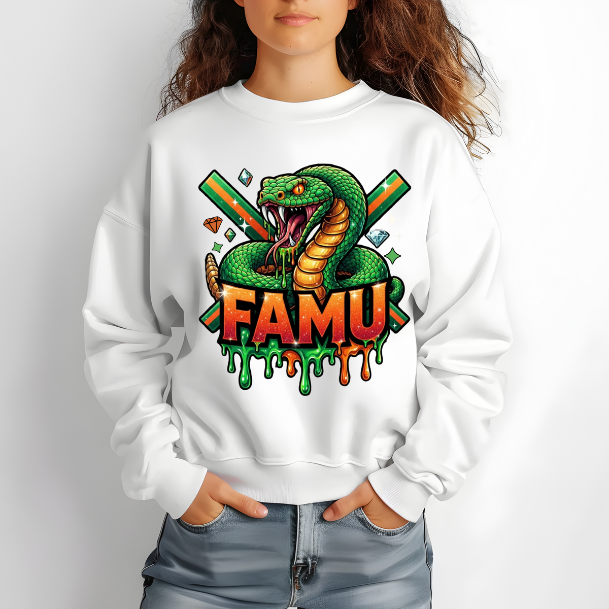 A woman wearing a white crewneck sweatshirt with a colorful graphic of a green snake, crossed green and orange guns, and the text 'FAMU' in dripping orange and green letters. She is wearing blue jeans against a light gray background.