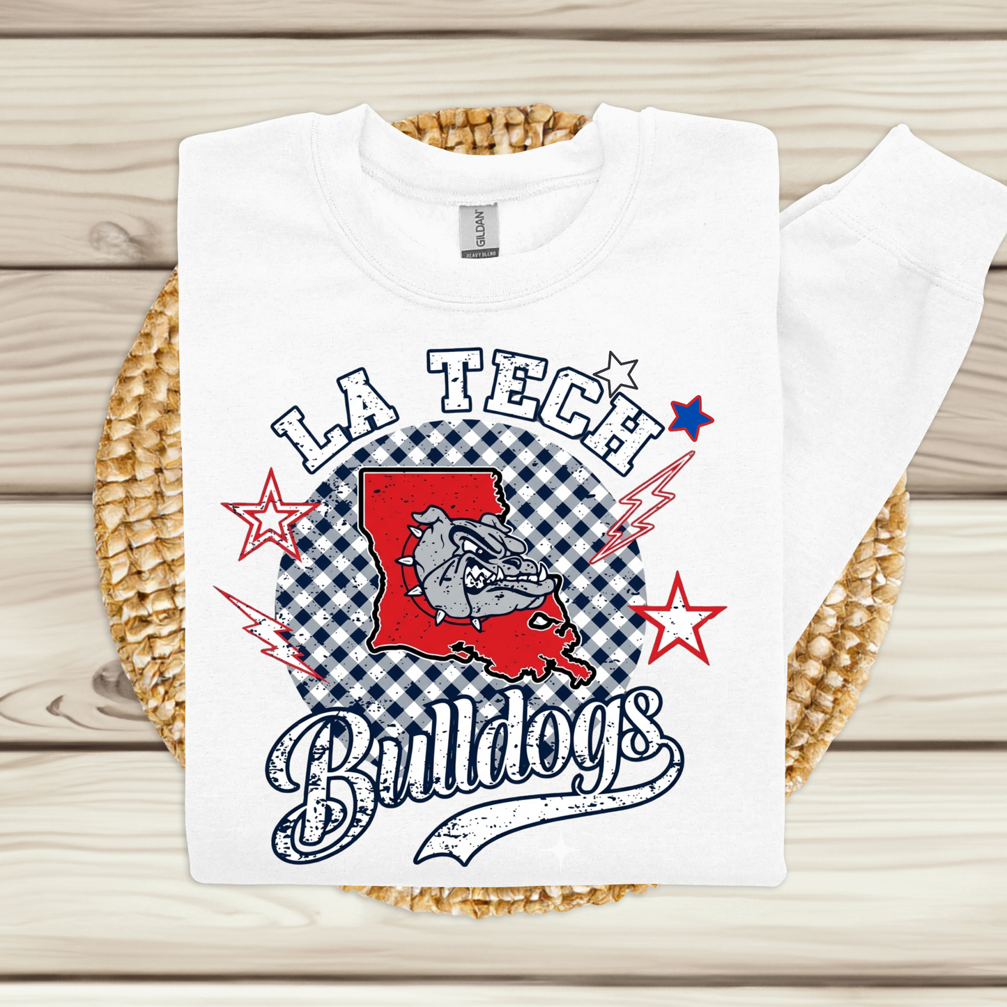 A white long-sleeve t-shirt with a 'LA Tech Bulldogs' graphic print, laid flat on a woven straw mat over a light wood plank background.