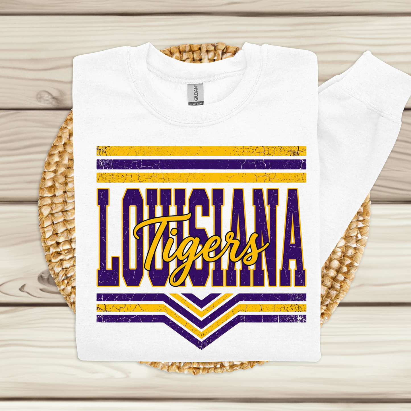 A white sweatshirt laid flat on a woven round mat over a light wood plank surface. The sweatshirt has a large front graphic reading "LOUISIANA Tigers" in purple and yellow with striped accents.