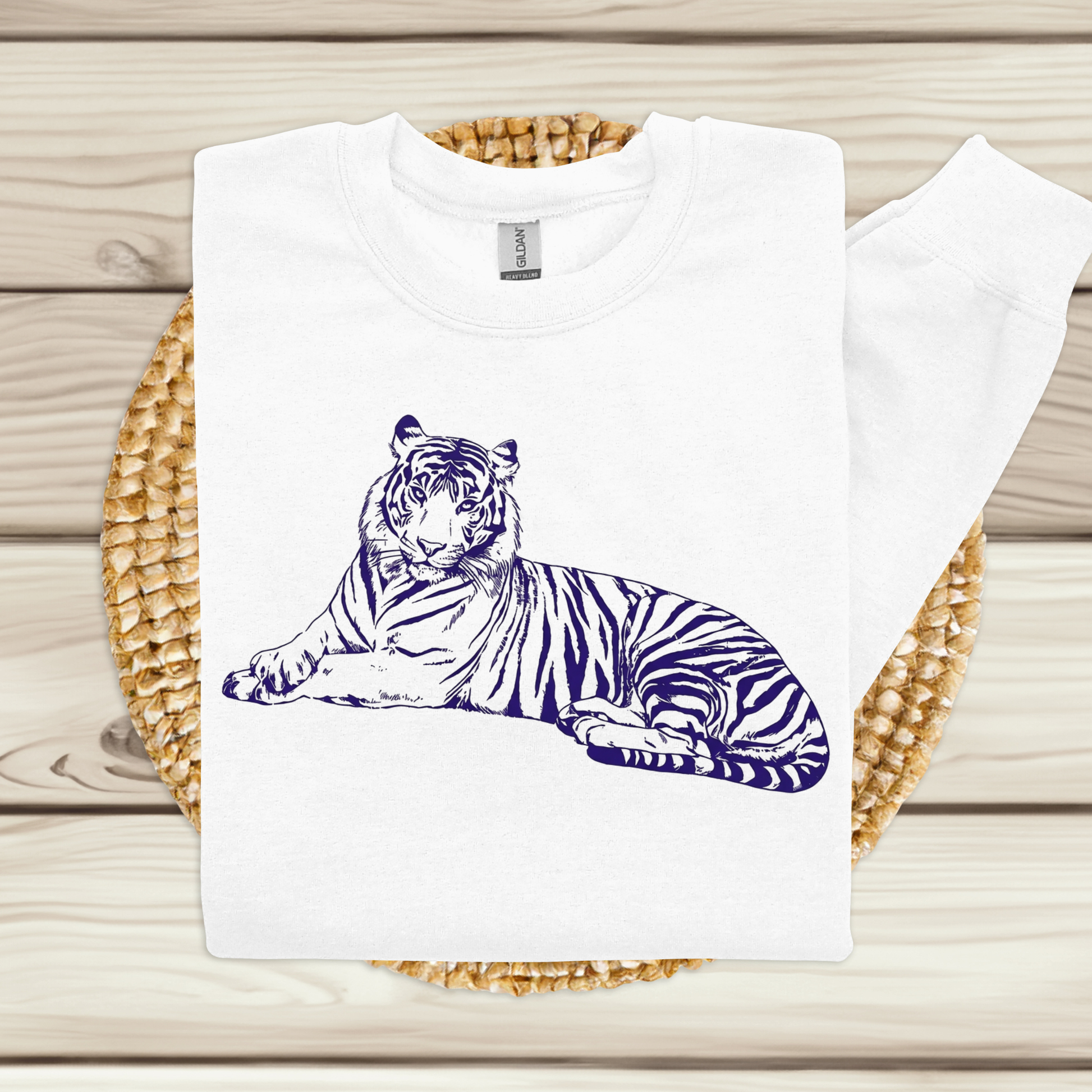 A white crewneck sweatshirt featuring a detailed navy blue line art illustration of a resting tiger on the front, displayed flat on a woven straw mat over a light wood plank background.