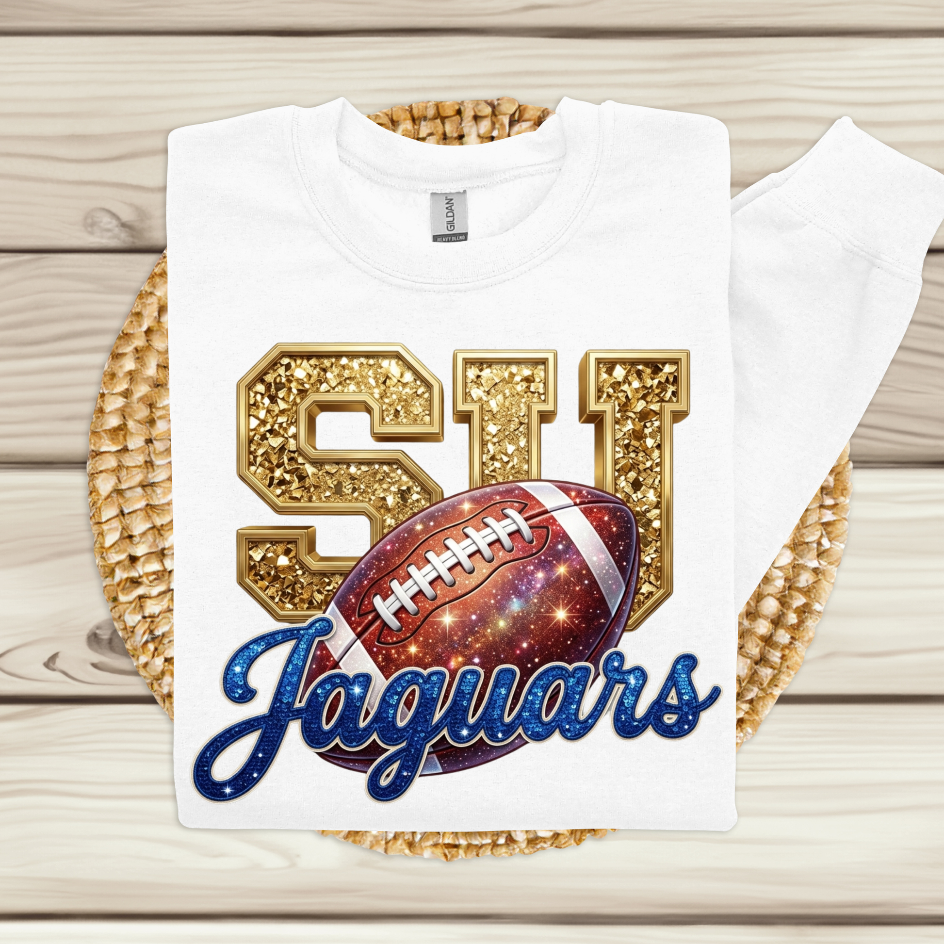A white short-sleeve t-shirt with a custom graphic print featuring glittery gold 'SI', a red and blue galaxy football, and blue cursive 'Jaguars' text, displayed on a woven placemat over a light wood background.
