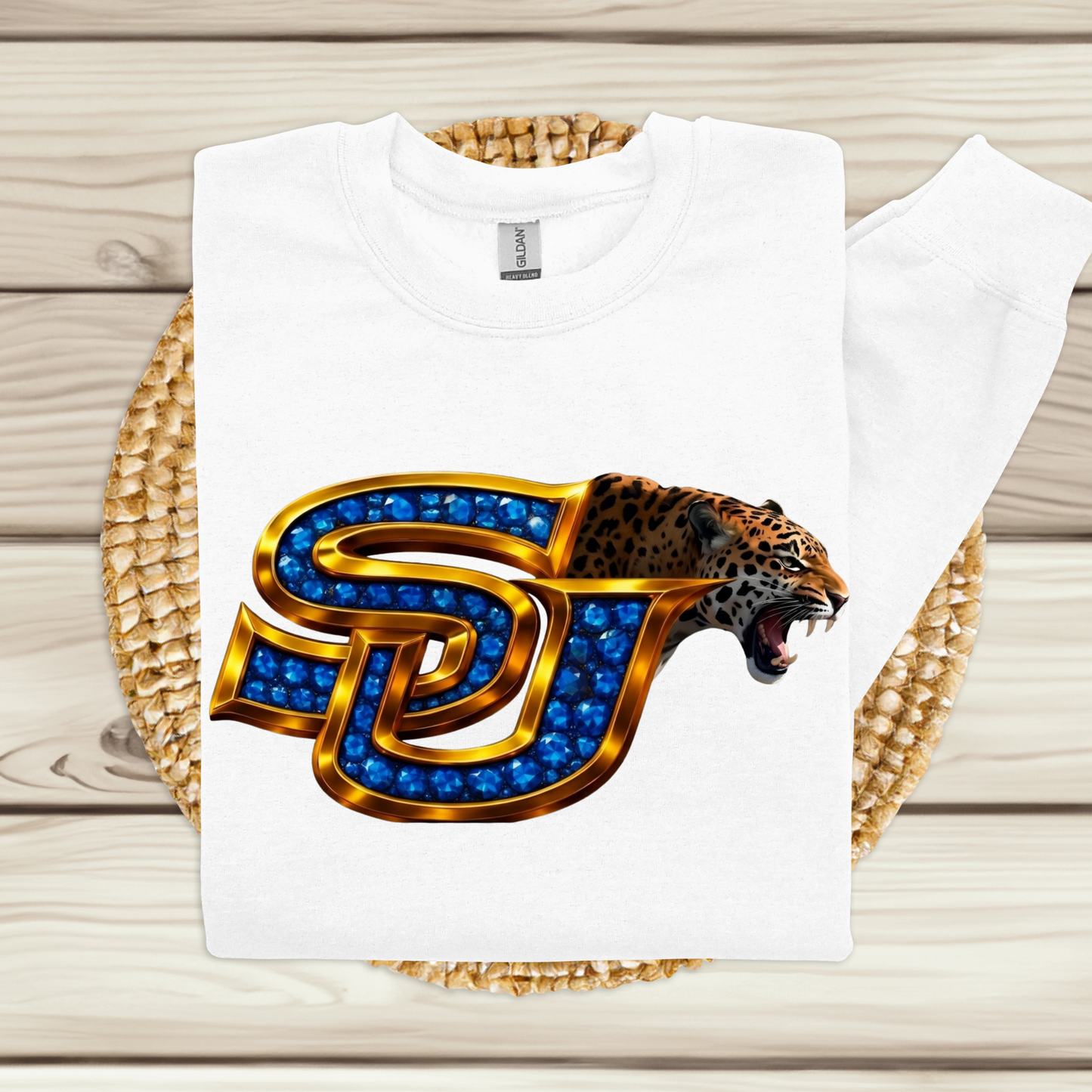 A white sweatshirt laid flat on a woven mat over a light wood surface, featuring a large front graphic of a roaring leopard head inside a stylized letter J outlined in gold and filled with blue gem-like details.