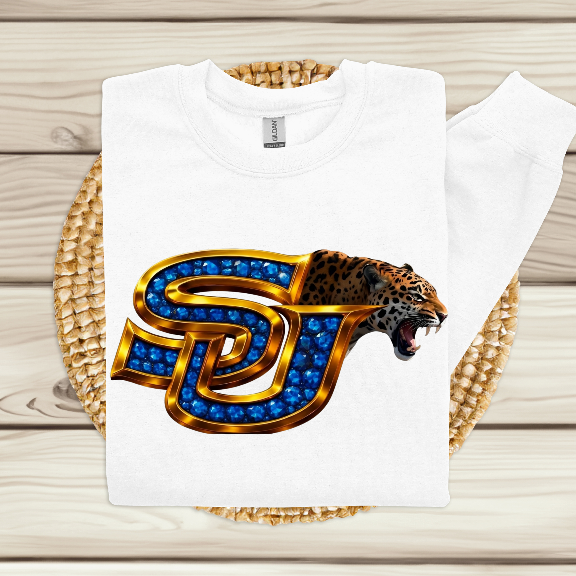 A white sweatshirt laid flat on a woven mat over a light wood surface, featuring a large front graphic of a roaring leopard head inside a stylized letter J outlined in gold and filled with blue gem-like details.