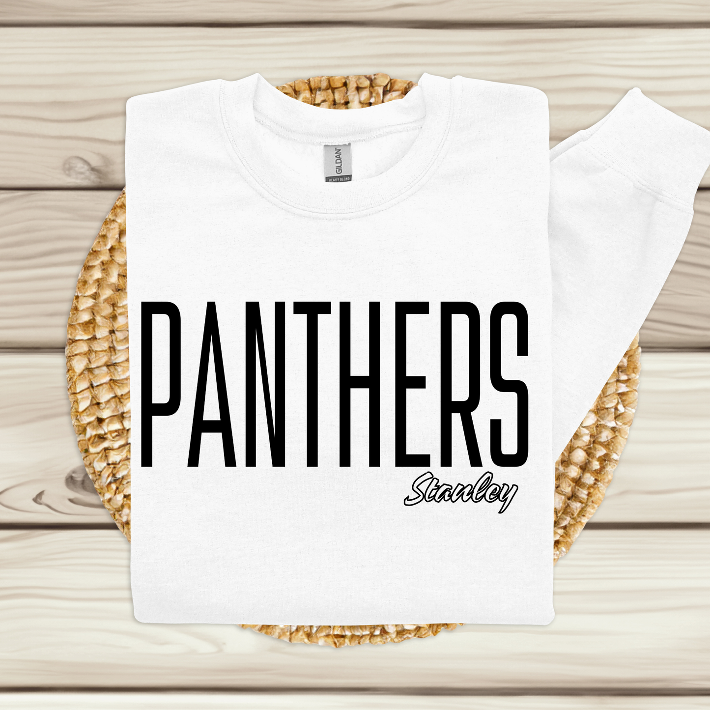 A white crewneck sweatshirt with 'PANTHERS' printed in bold black letters and 'Stanley' in script below, laid flat on a woven round mat over a light wood plank background.