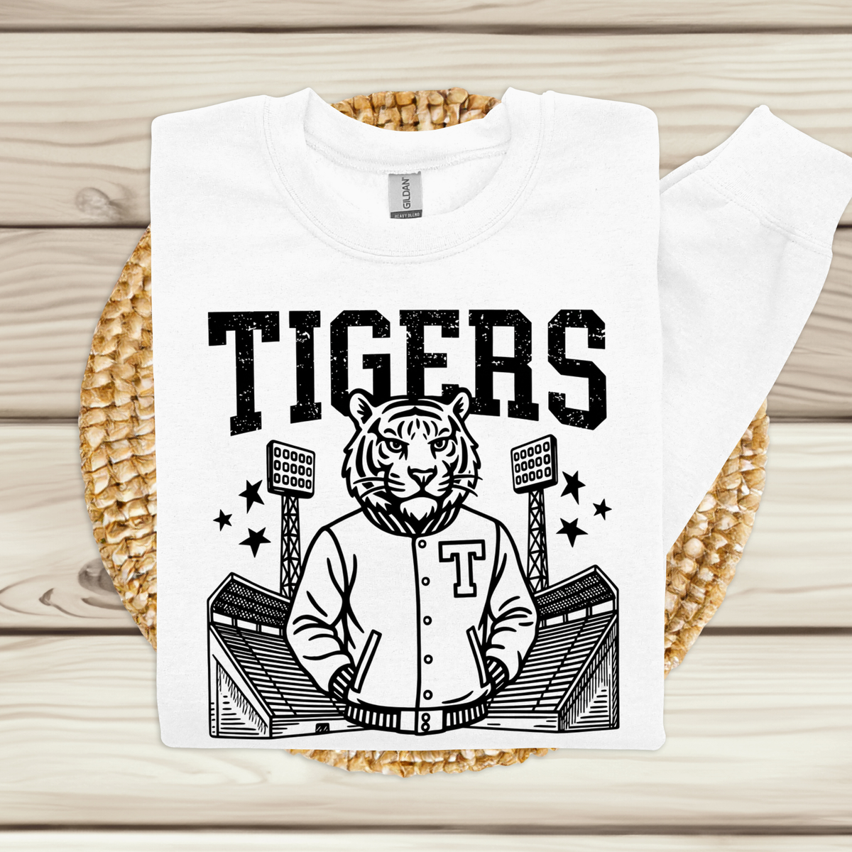 A white long-sleeve T-shirt laid flat on a woven round mat over a light wood plank surface. The shirt has a large black graphic print featuring the word "TIGERS" above a tiger mascot in a baseball jersey with a large "T" on the chest, stadium stands, and stars.