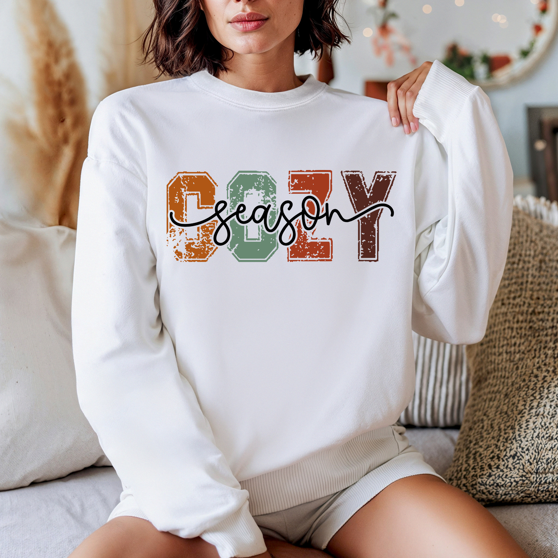 A woman wearing a white crewneck sweatshirt with a colorful distressed graphic that reads 'COZY season' in orange, green, and brown block letters with black script. She is sitting on a couch in a cozy, warm-toned living room setting.
