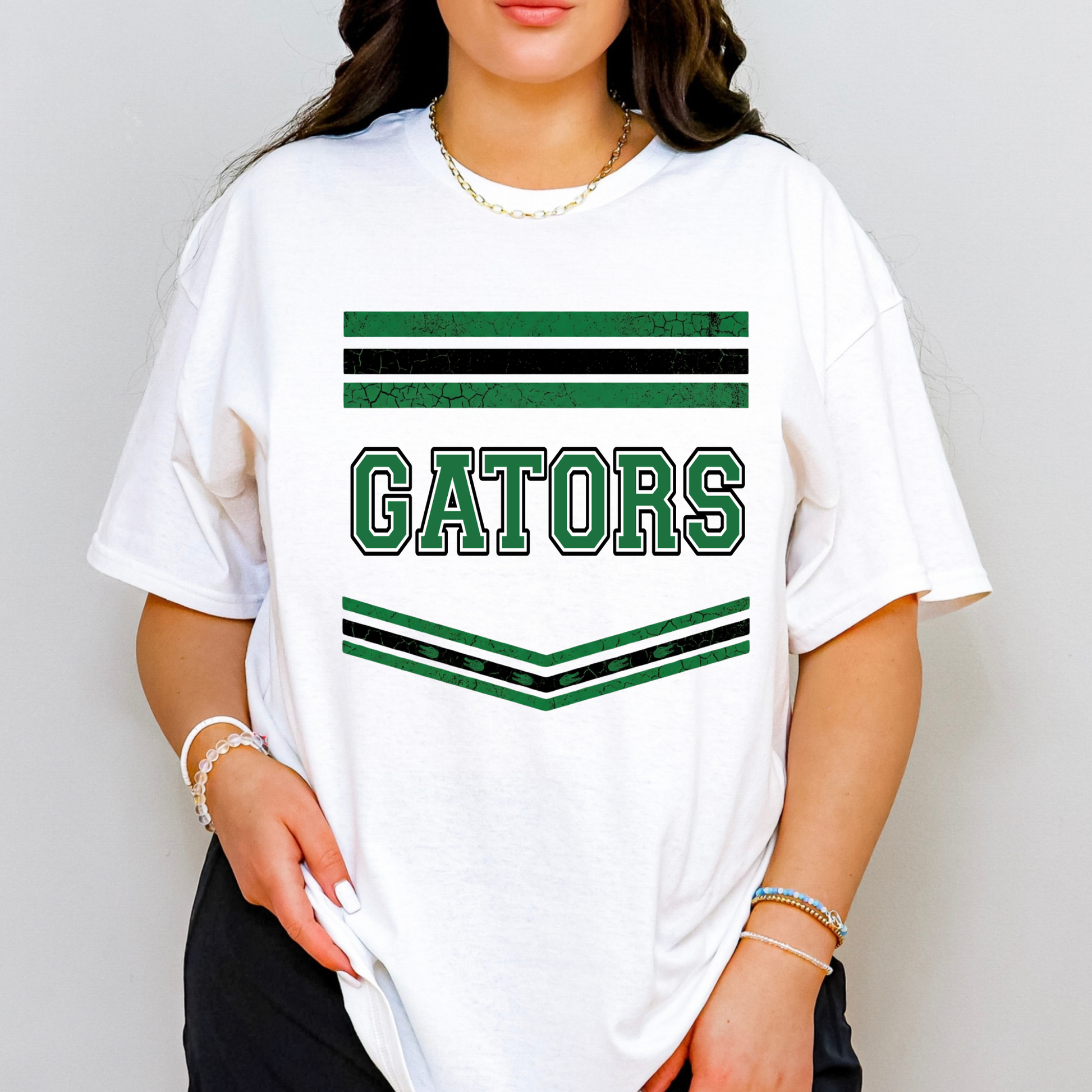 A woman wearing a white short-sleeve graphic T-shirt with green and black stripes and the word 'GATORS' across the chest, photographed against a light gray studio background.