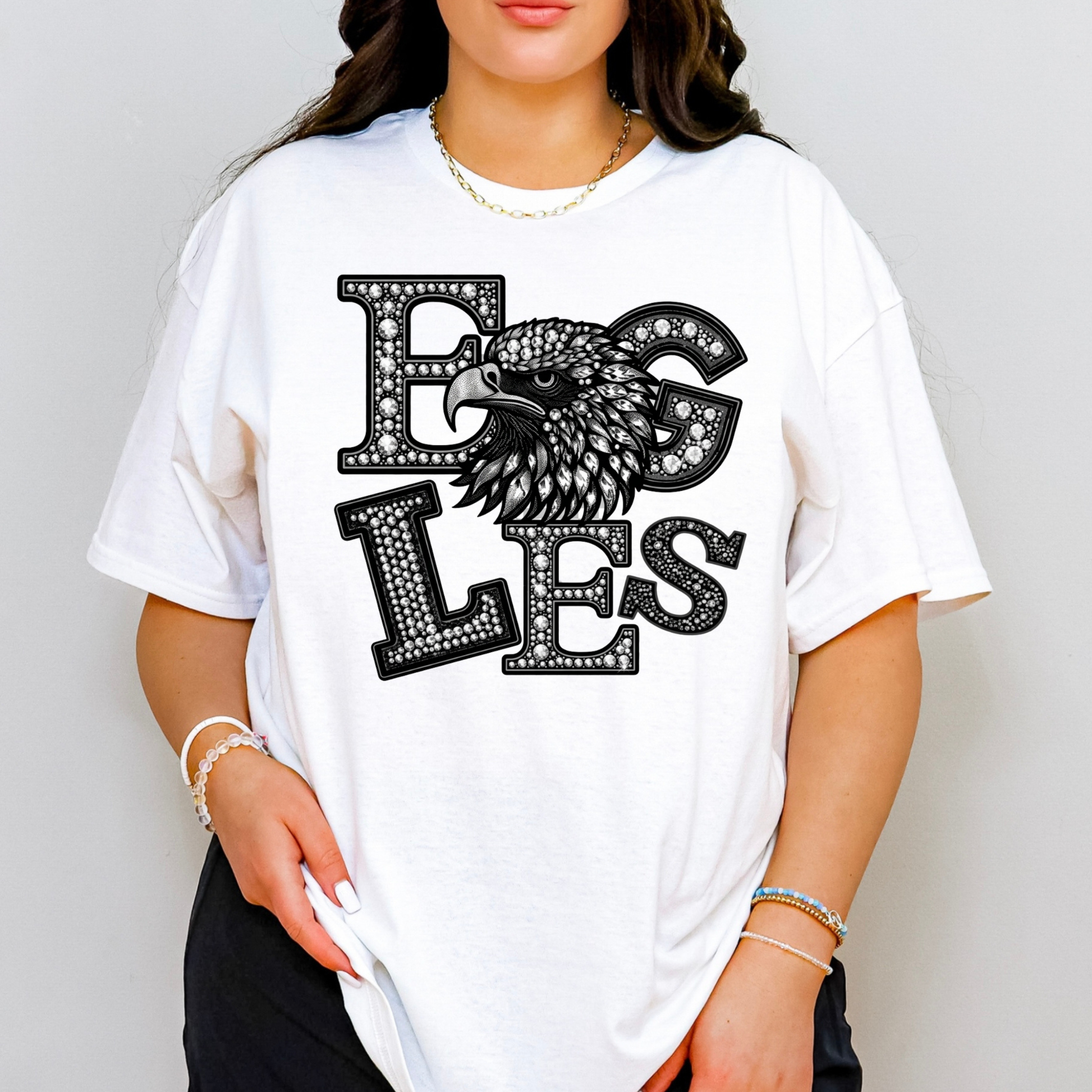 A woman wearing a white oversized t-shirt with a black graphic that reads 'EGS LES' in a studded font, featuring an eagle head design. The background is a plain light gray wall.