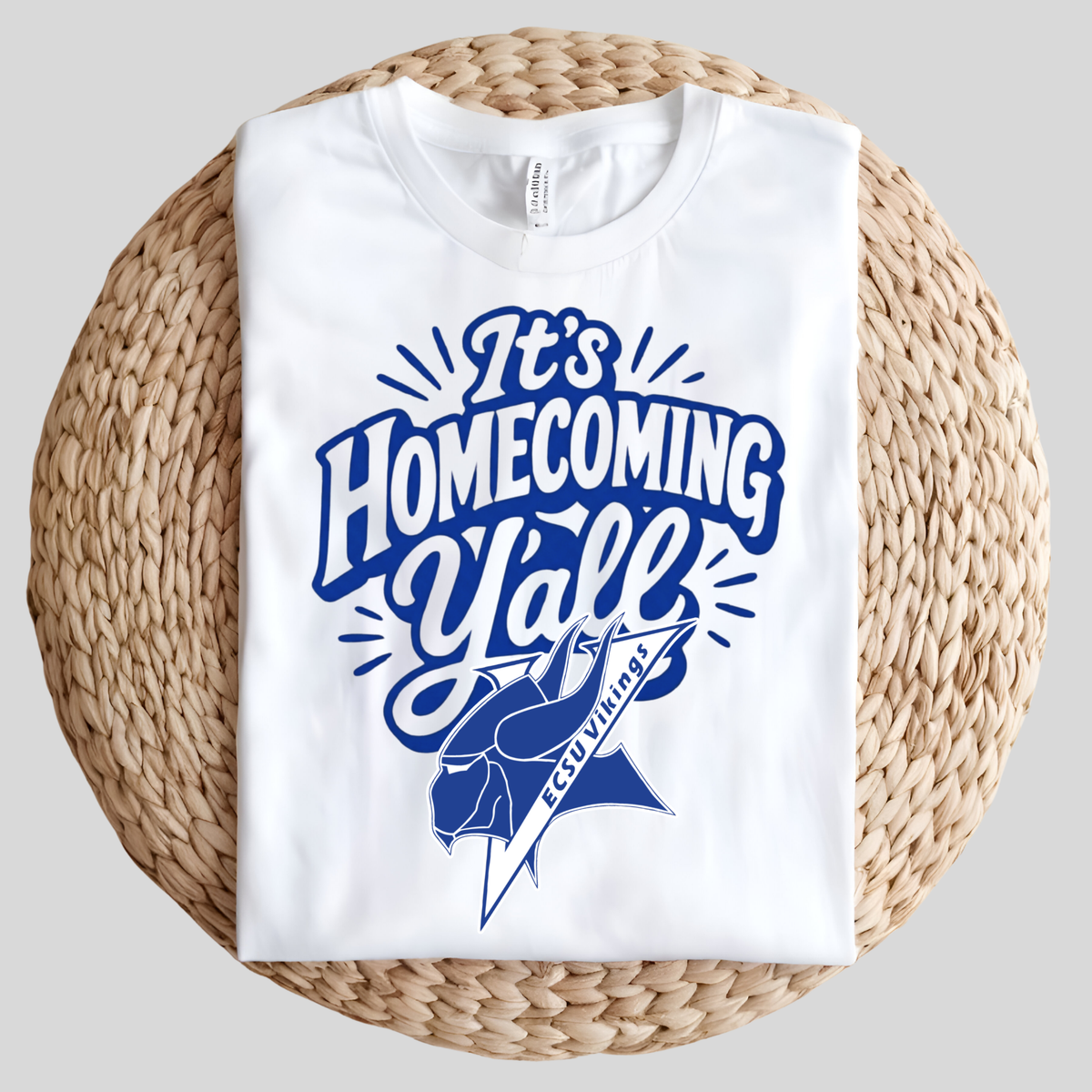 A folded white t-shirt with a blue 'It's Homecoming Yell' graphic and a Spartan helmet design, displayed on a round woven straw mat against a neutral gray background.