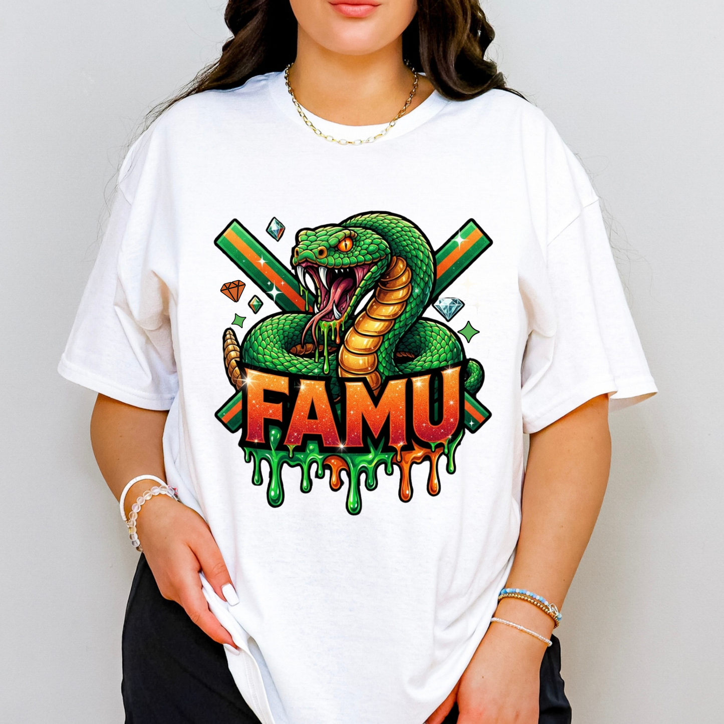 A woman wearing a white oversized graphic t-shirt featuring a green snake coiled around the word 'FAMU' in dripping orange and green letters, with crossed green and orange bars in the background.