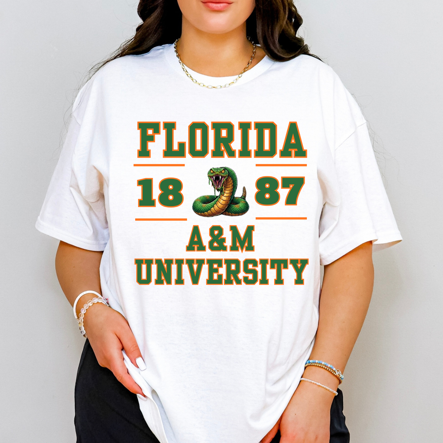 A woman wearing a white oversized graphic T-shirt with green and orange text reading 'FLORIDA 1887 A&M UNIVERSITY' and a snake graphic on the front, photographed against a light gray studio background.
