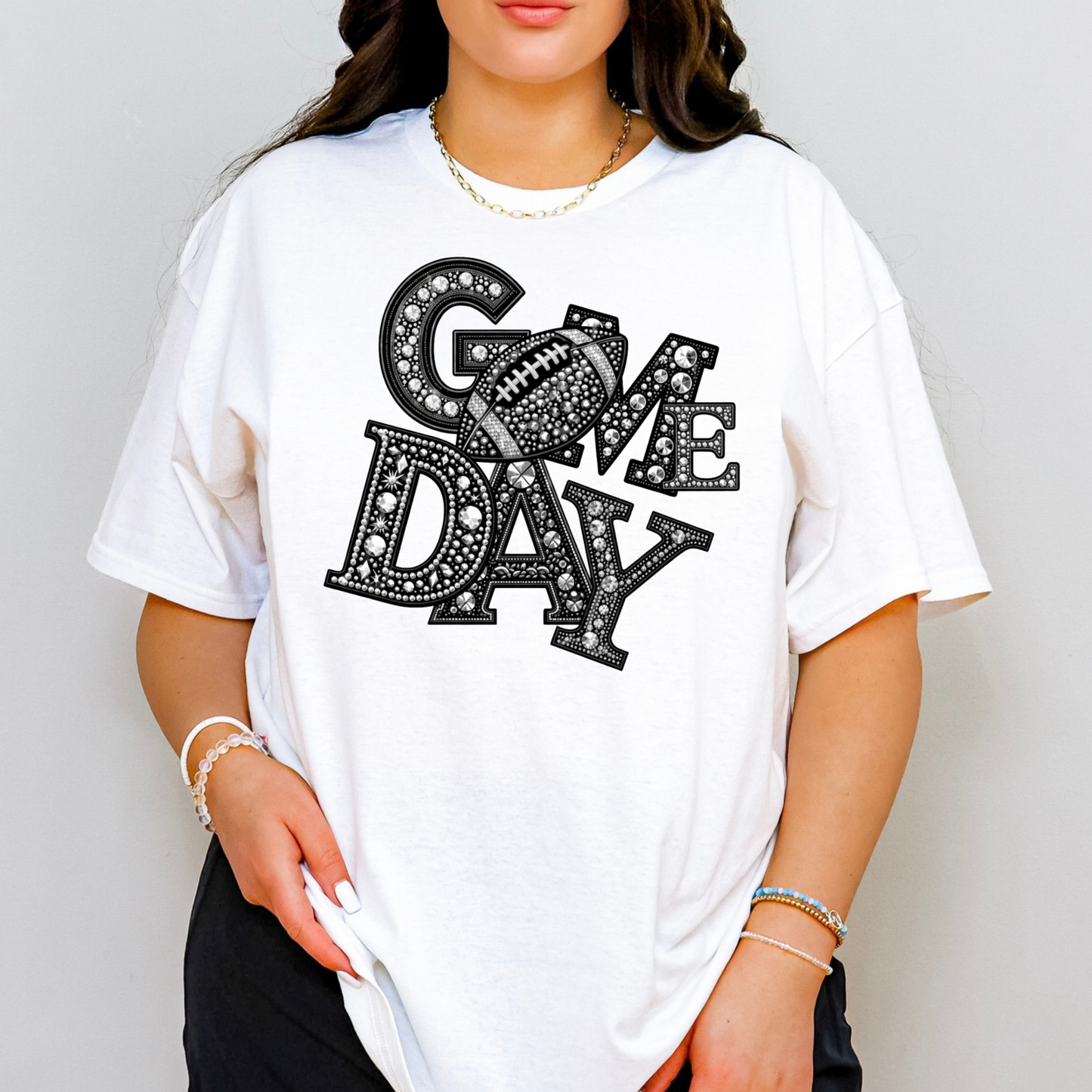 A woman wearing an oversized white graphic T-shirt with the words 'GAME DAY' in black letters filled with rhinestone-like patterns. The photo is cropped from the lower face to the hips against a plain light gray background.
