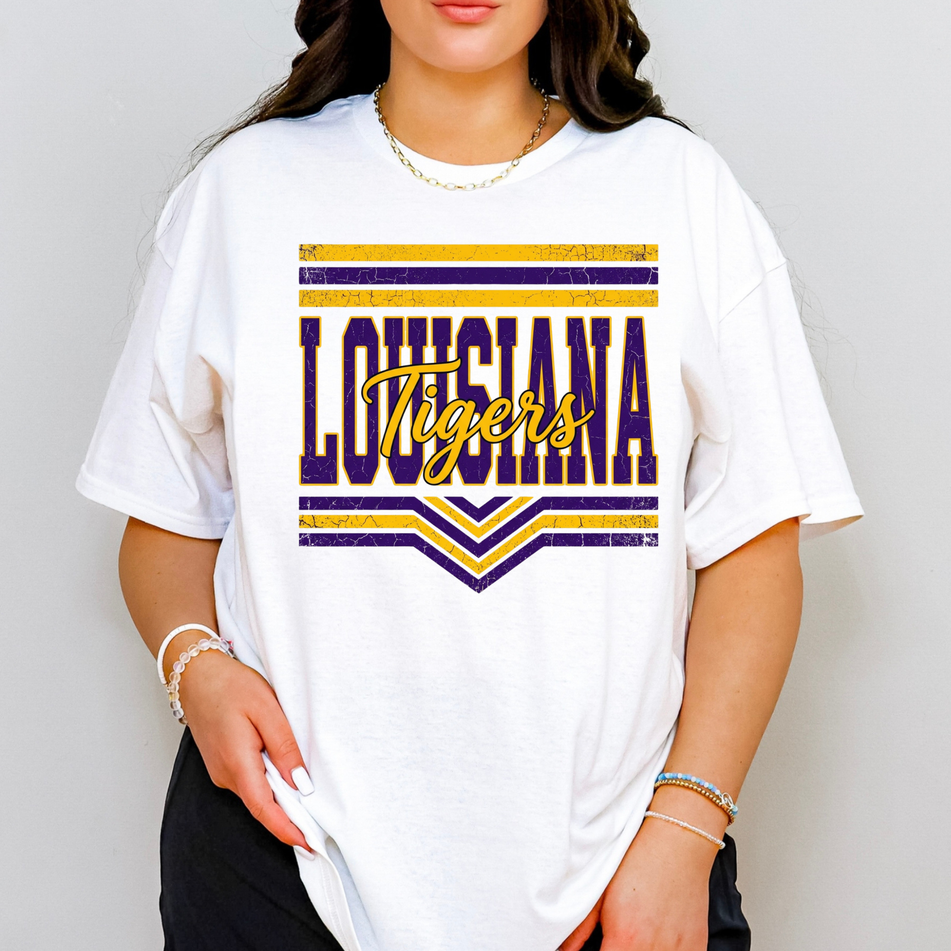 A woman wearing a white oversized graphic T-shirt with a distressed purple and gold 'LOUISIANA Tigers' design on the front, paired with black pants and gold jewelry, against a light gray background.