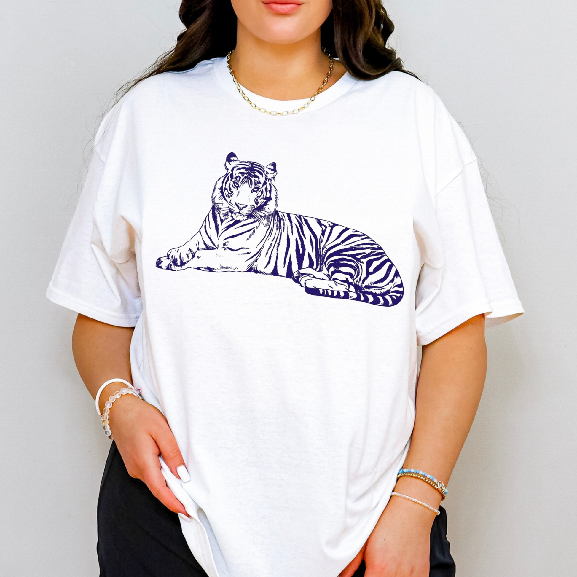 A woman wearing a white oversized short-sleeve t-shirt with a navy blue line art graphic of a lying tiger on the chest, paired with black pants and gold jewelry, against a light gray background.