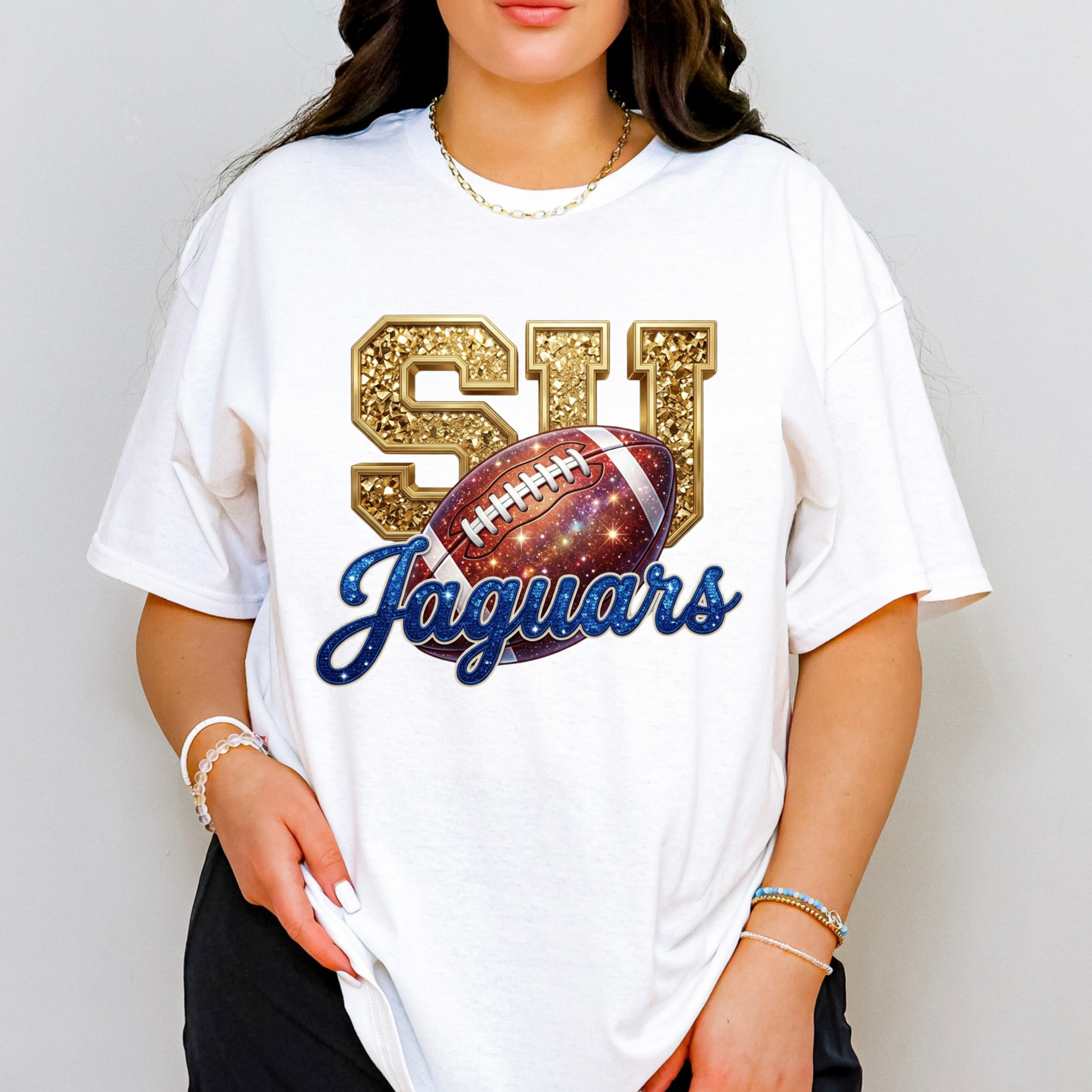 A woman wearing a white oversized graphic T-shirt with a gold glittery 'SU' lettering, a football graphic, and 'Jaguars' in blue script, paired with black pants and gold jewelry against a light gray background.