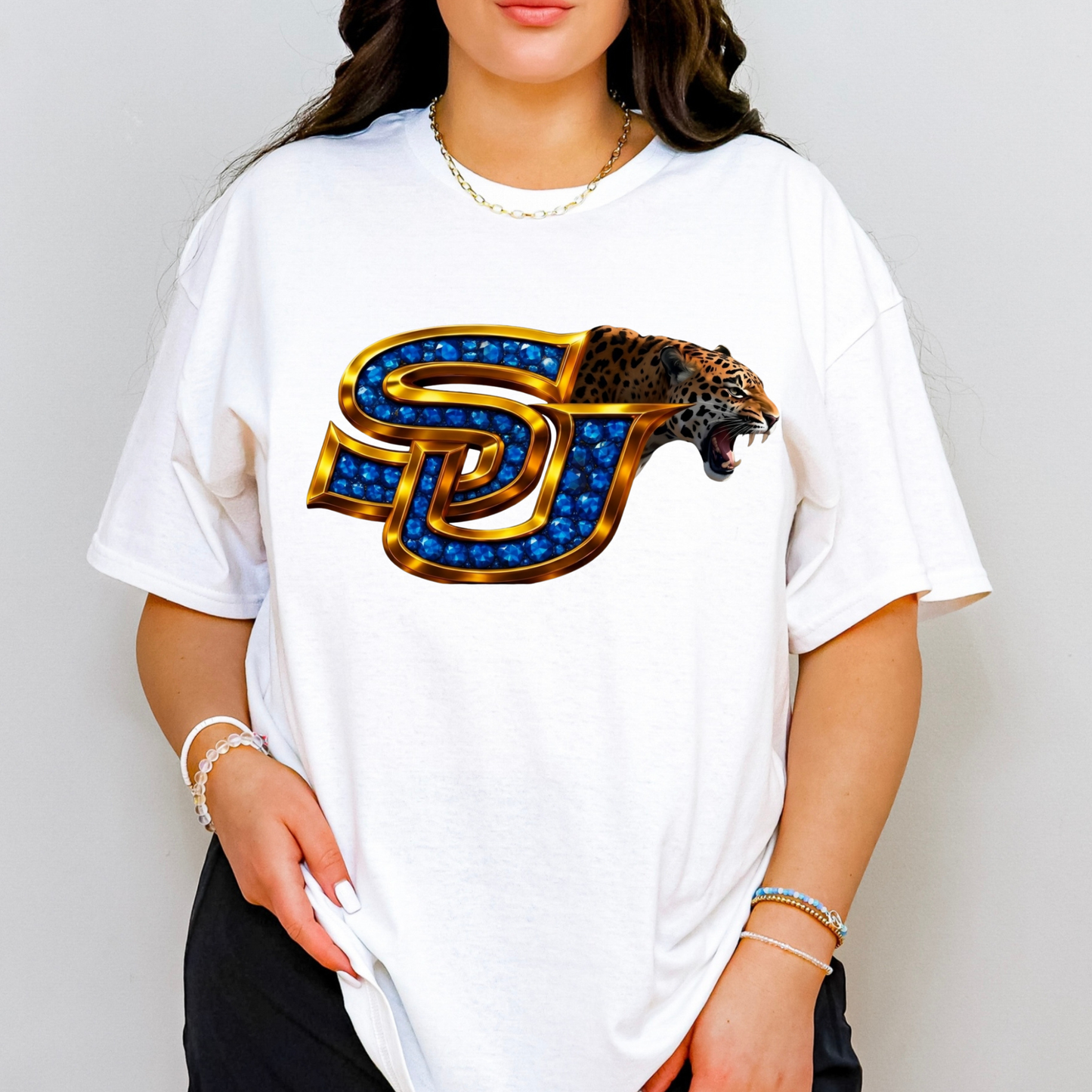 A woman wearing a white oversized t-shirt with a large graphic of the 'SJ' Utah State University logo, styled with blue gem details, gold outlines, and a leopard head on the right side. The background is a clean, neutral gray studio wall.
