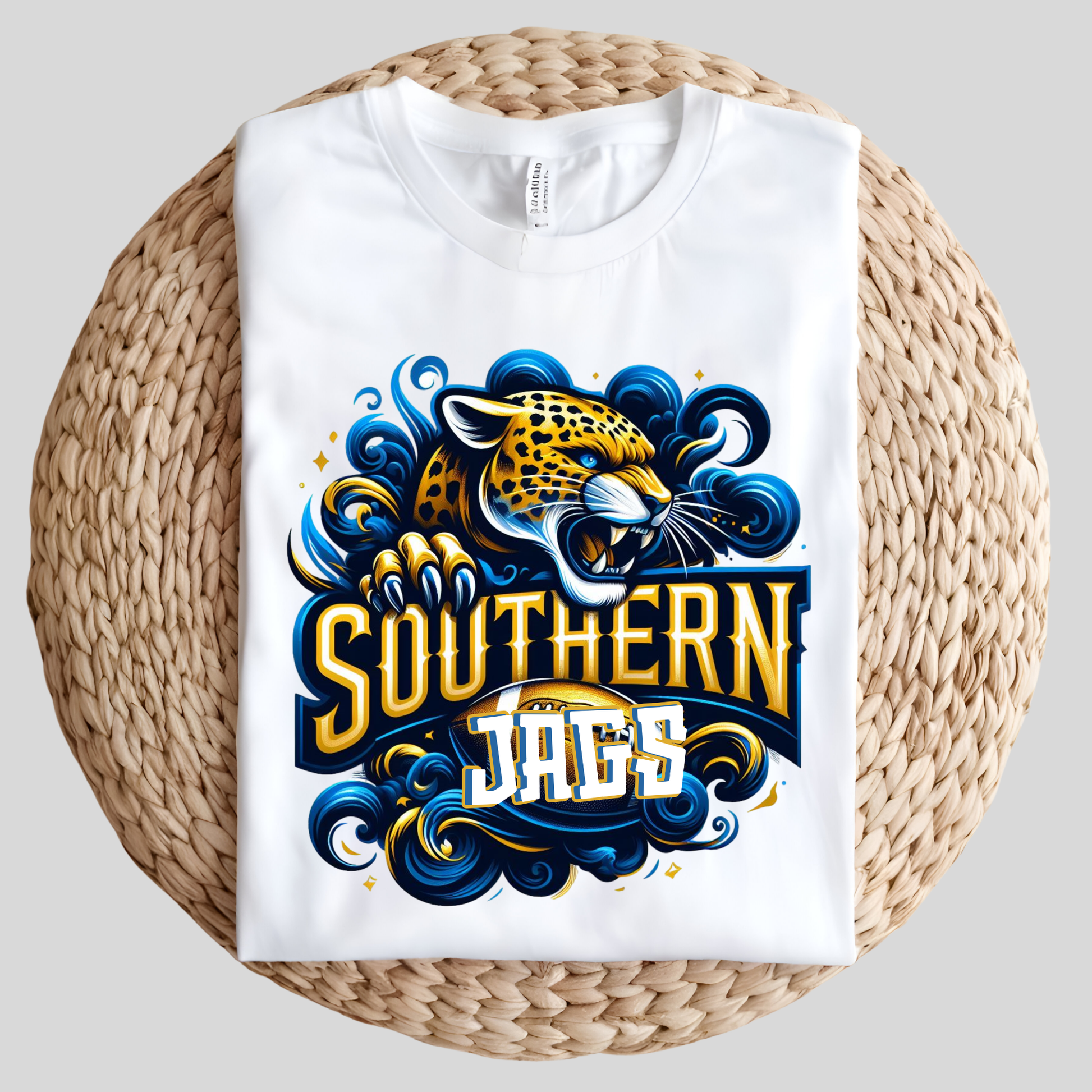 A white short-sleeve T-shirt folded flat on a round woven straw mat, featuring a large graphic print of a roaring jaguar head and the words 'SOUTHERN JAGS' in bold yellow, blue, and white lettering with decorative swirls.