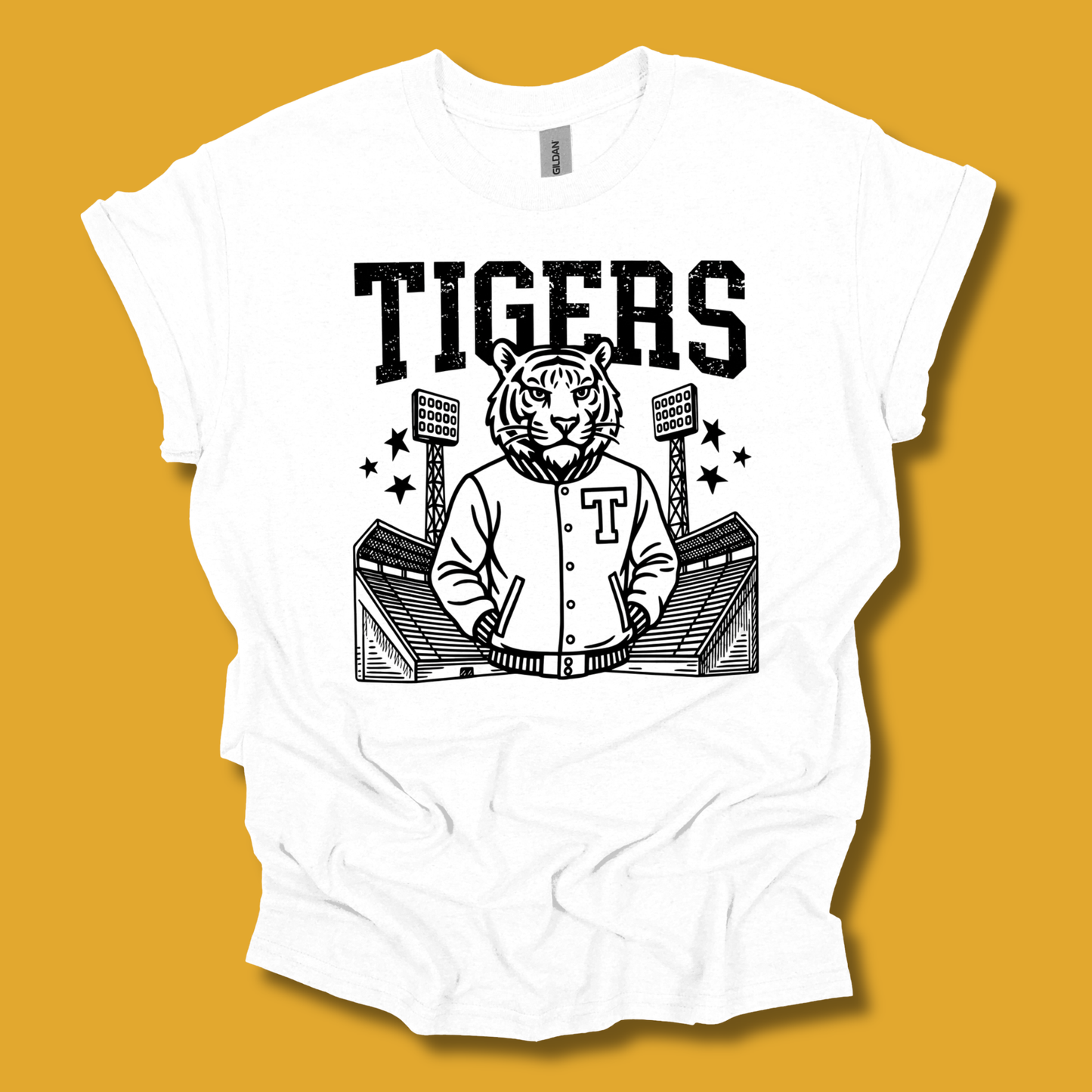 A white short-sleeve t-shirt with a black graphic print of a tiger mascot wearing a varsity jacket in front of a stadium, with the word 'TIGERS' printed above. The shirt is displayed flat against a solid mustard yellow background.