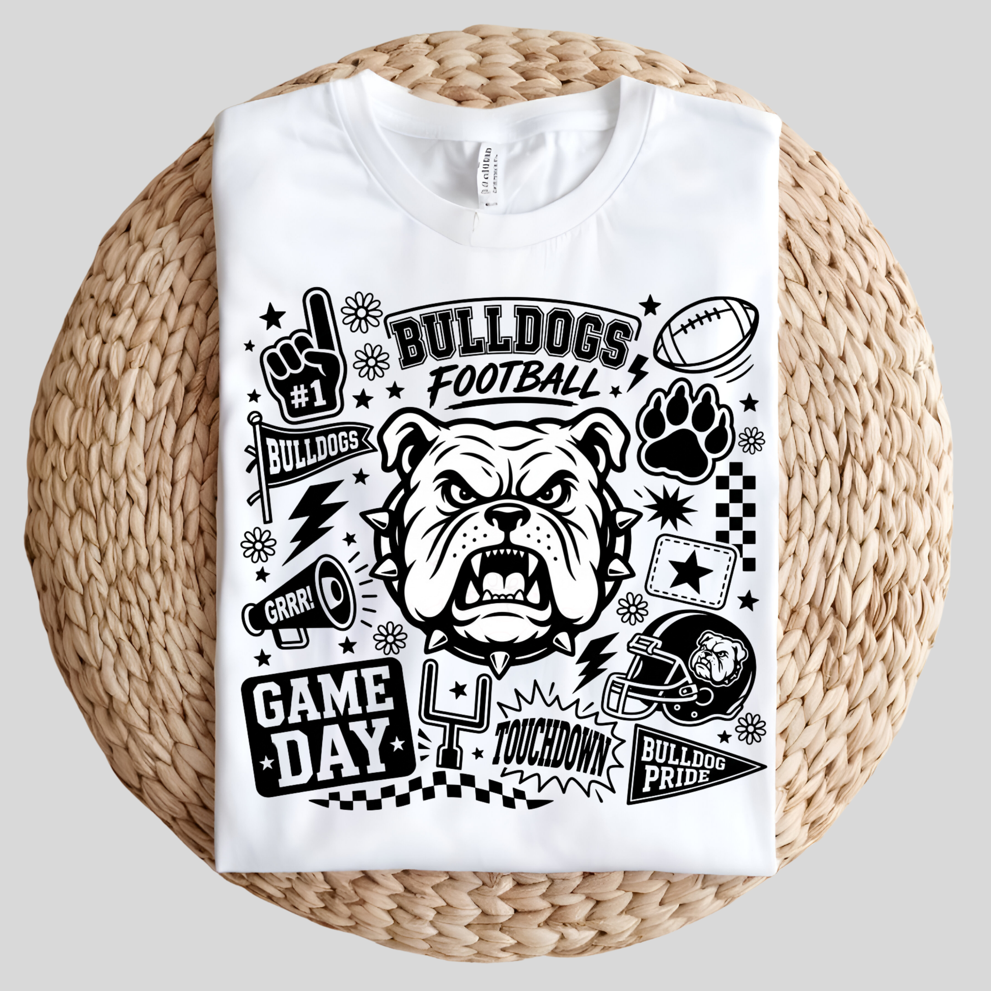 A white short-sleeve t-shirt with a black graphic print of an angry bulldog and football-themed designs, displayed flat on a round woven straw mat against a light gray background.