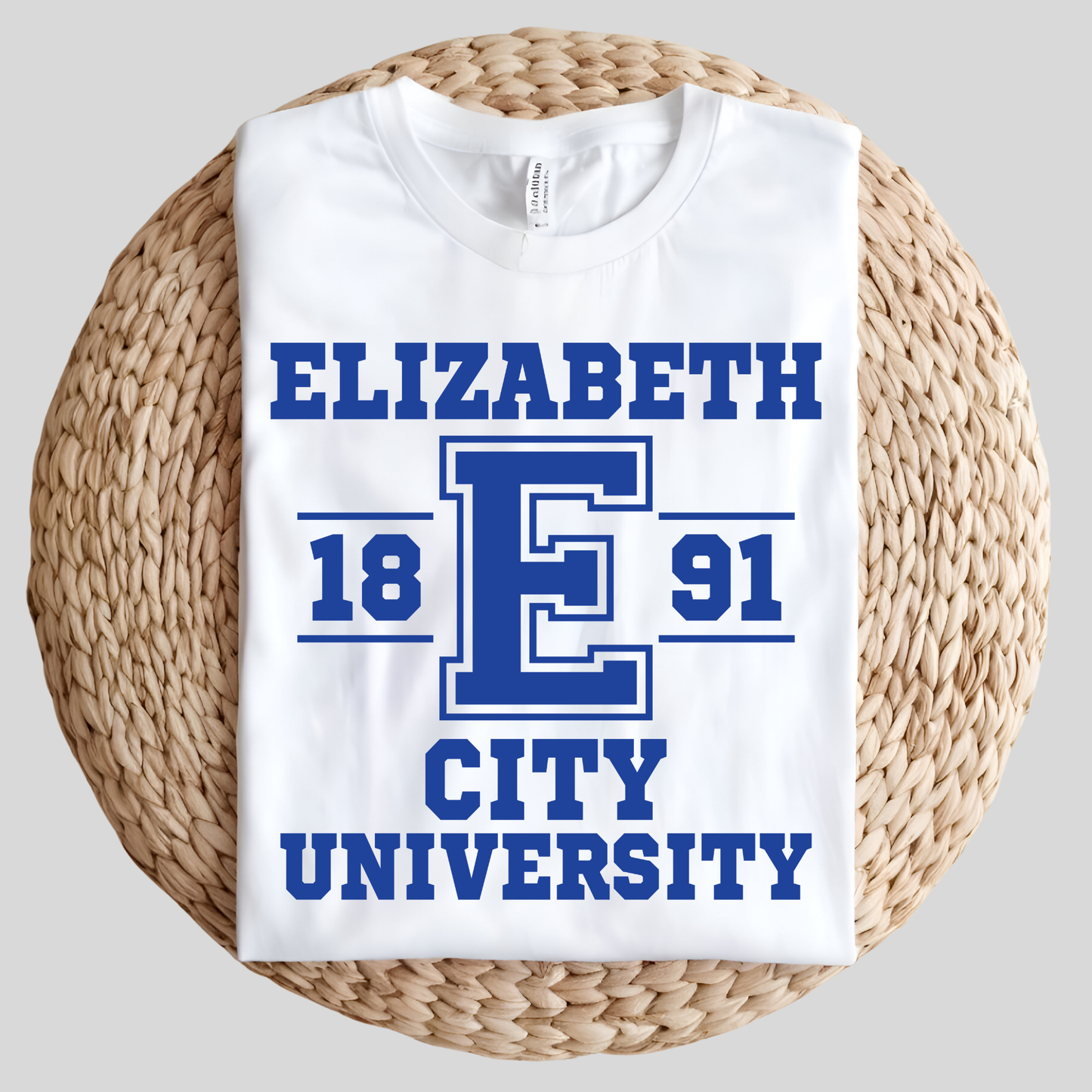 A white short-sleeve T-shirt folded and displayed on a round woven straw mat against a light gray background. The shirt has a bold blue graphic reading 'ELIZABETH E 1891 CITY UNIVERSITY' with a large block letter E in the center.