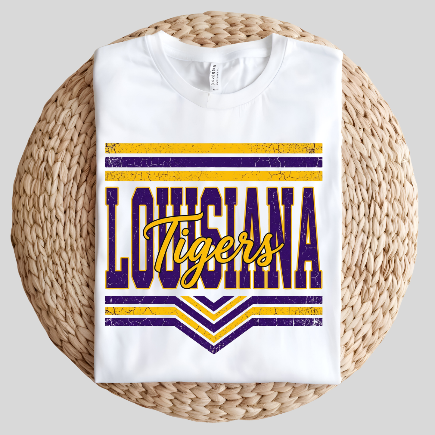 A white short-sleeve T-shirt with a distressed purple and yellow 'Louisiana Tigers' graphic, displayed flat on a round woven straw mat against a light gray background.