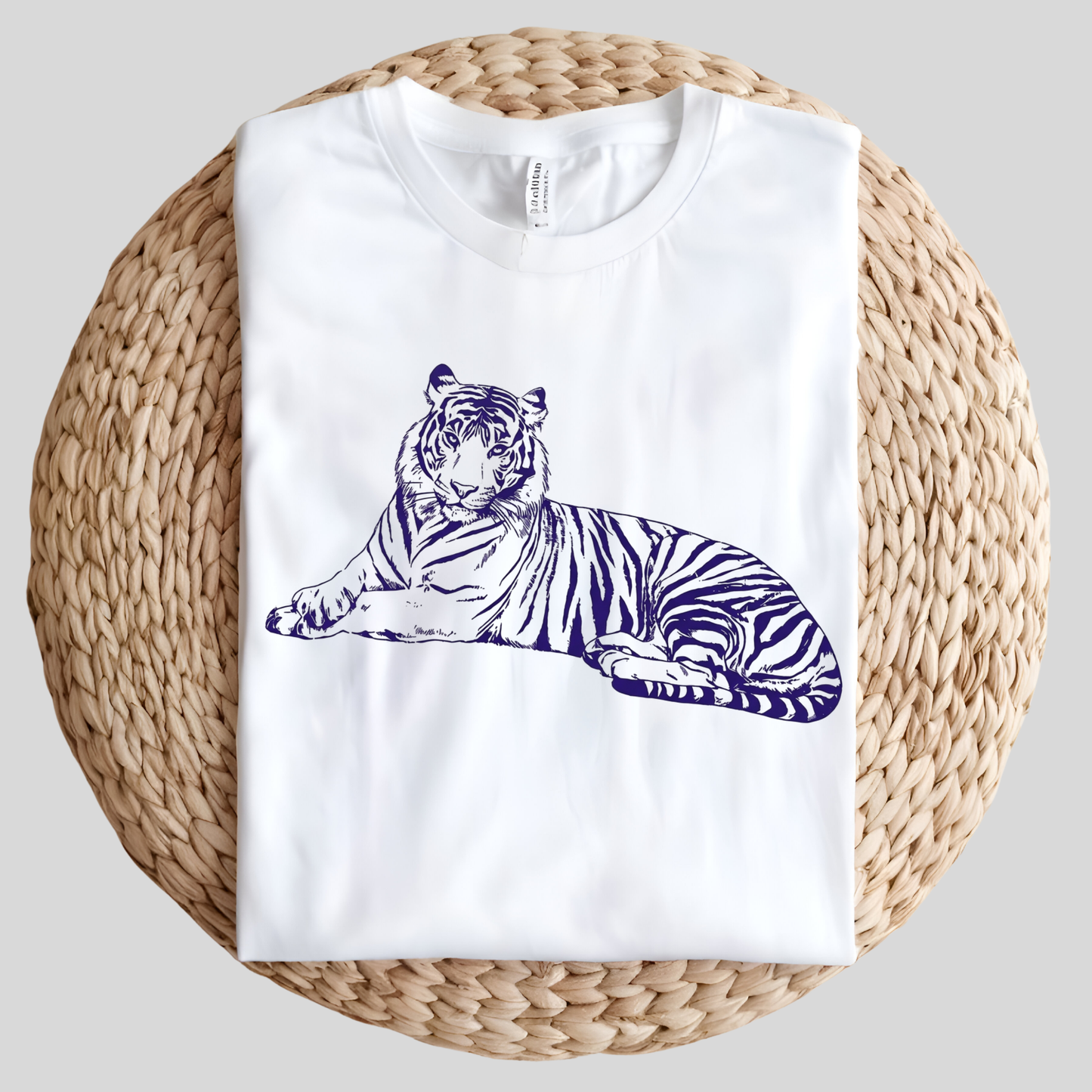 A folded white crewneck t-shirt featuring a dark blue line art illustration of a resting tiger, displayed on a round woven straw mat against a neutral gray background.