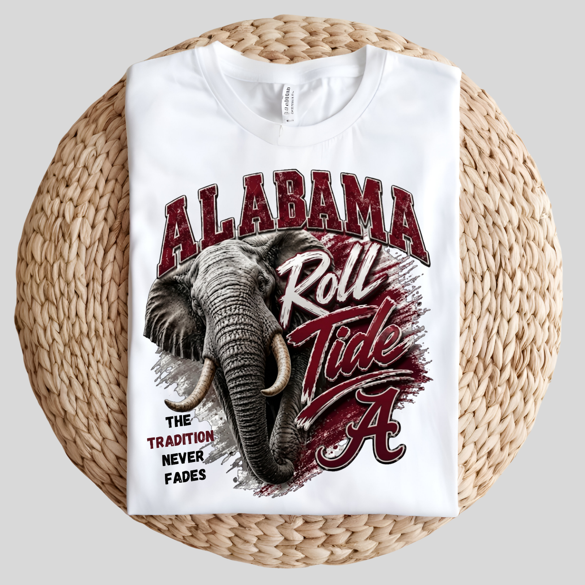 A folded white graphic T-shirt displayed on a round woven straw mat against a light gray background. The shirt features a large printed design with the words 'ALABAMA Roll Tide' and 'The Tradition Never Fades,' along with an elephant illustration and the Alabama 'A' logo.
