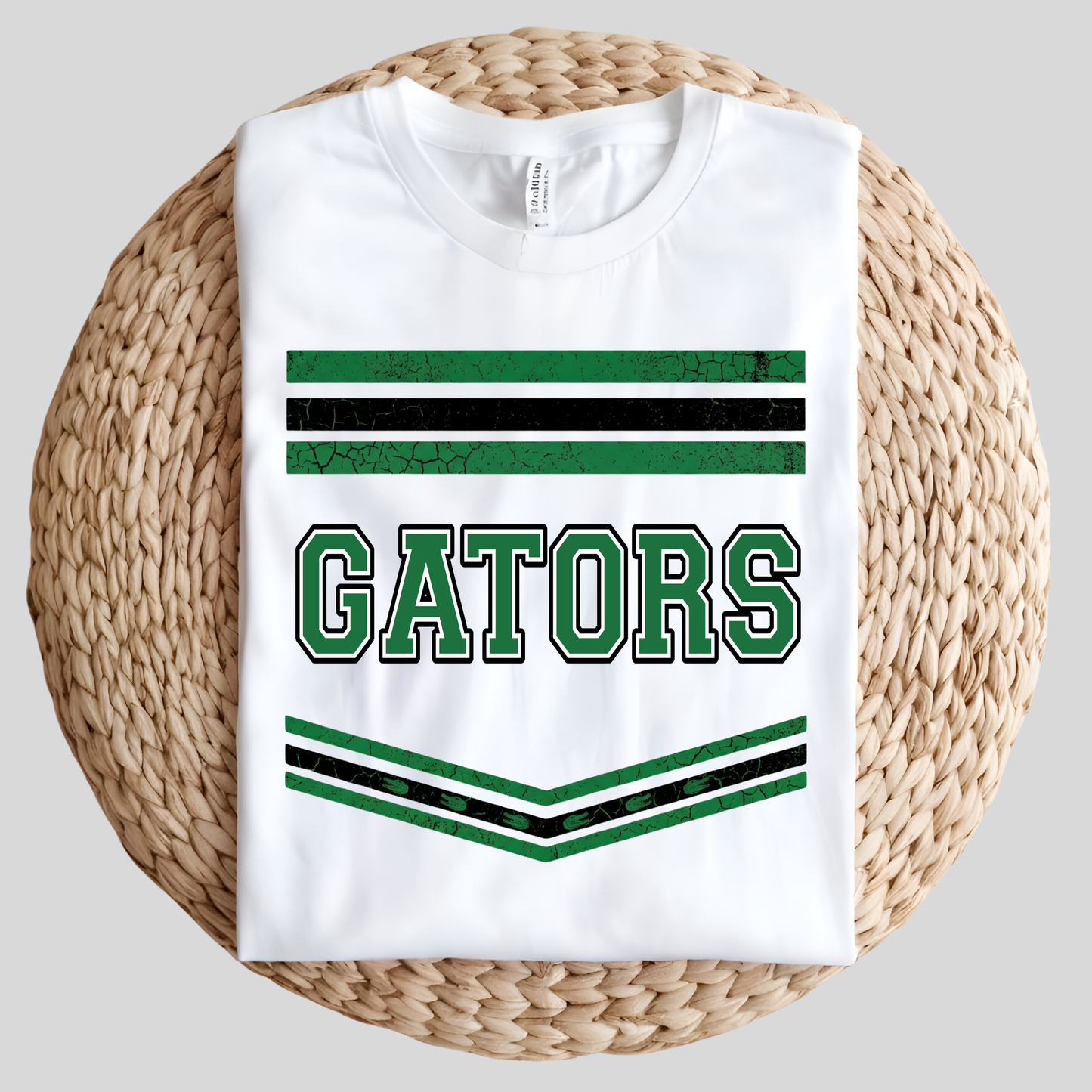 A white short-sleeve t-shirt with a green and black 'GATORS' collegiate graphic, laid flat on a round woven straw mat against a light gray background.