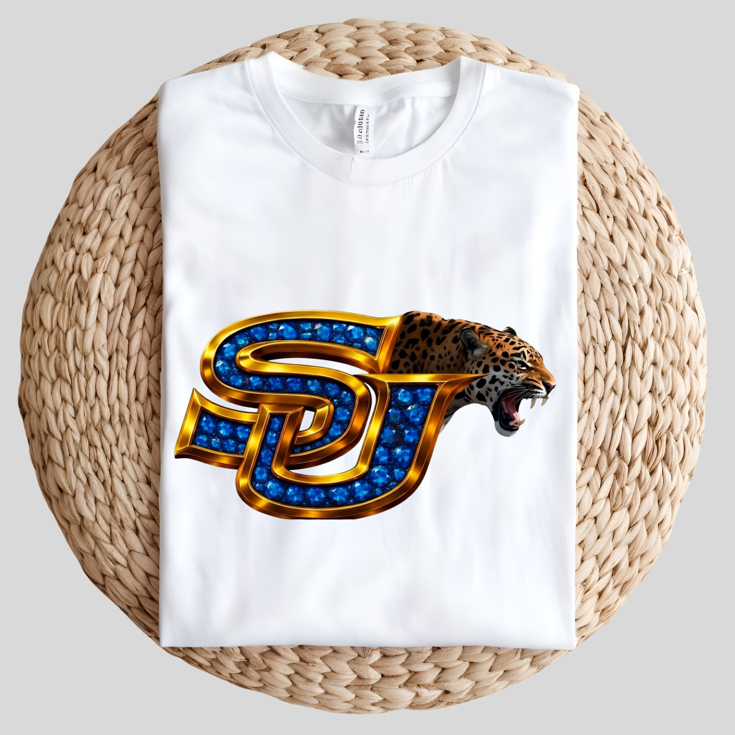 A folded white t-shirt displayed on a round woven straw mat against a light gray background. The t-shirt features a graphic of a roaring leopard head combined with a stylized 'SJ' monogram filled with blue gem-like details and outlined in gold.