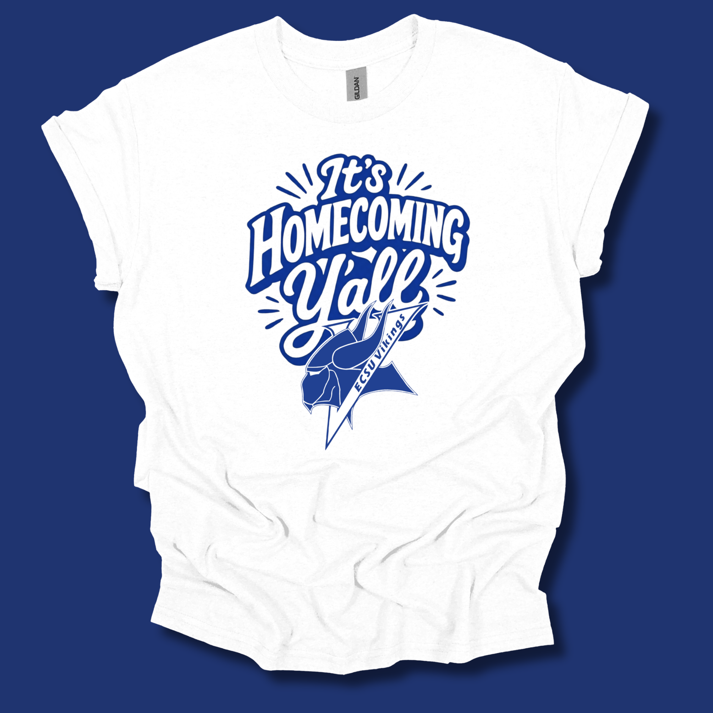 A white short-sleeve t-shirt with a blue graphic print reading 'It's Homecoming Y'all' and a football player silhouette, displayed against a solid dark blue background.