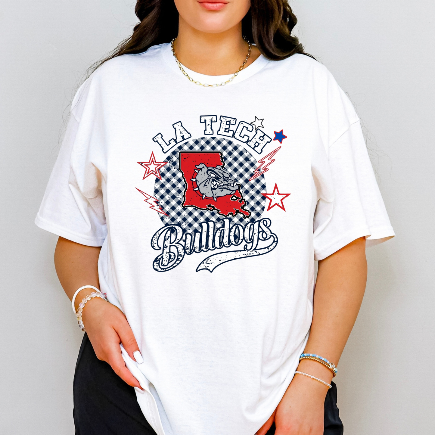 A woman wearing an oversized white graphic t-shirt featuring the 'LA Tech Bulldogs' logo in red, black, and blue, paired with black pants and gold jewelry, set against a plain light gray background.