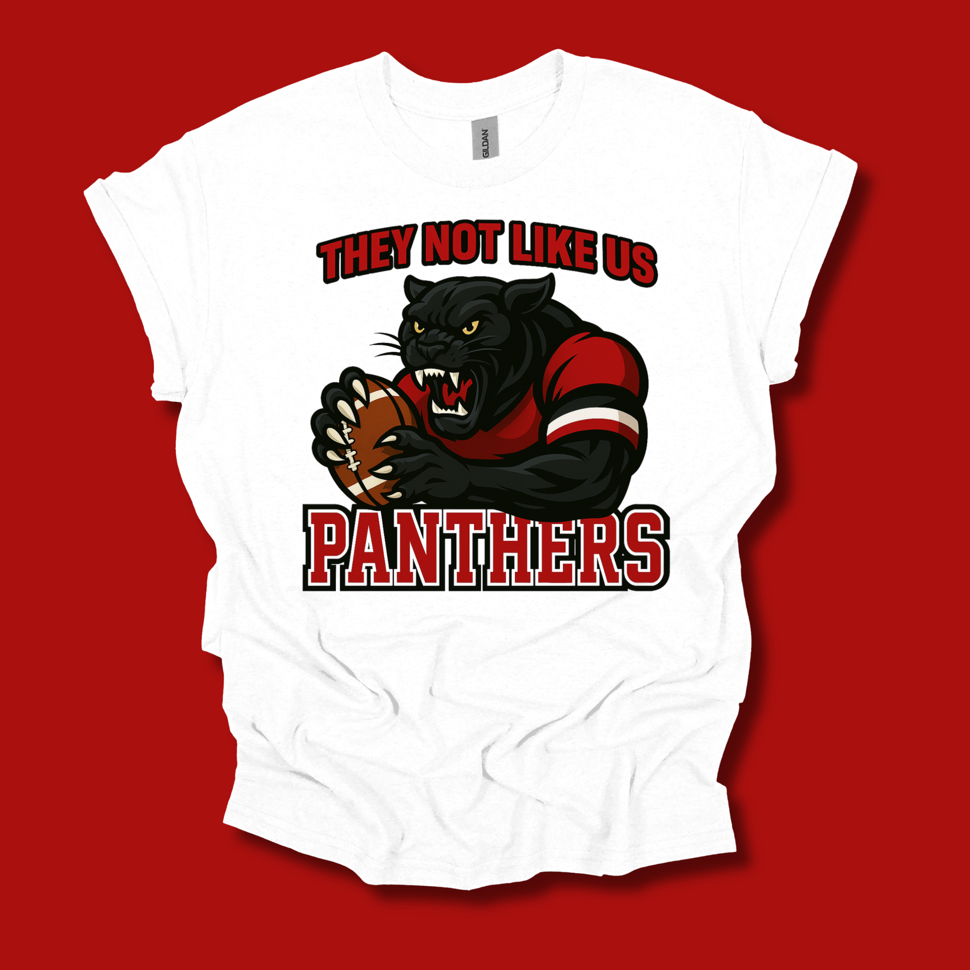A white short-sleeve graphic t-shirt featuring a cartoon panther mascot holding a football, with the text 'THEY NOT LIKE US PANTHERS' in red and black. The t-shirt is displayed flat against a solid red background.