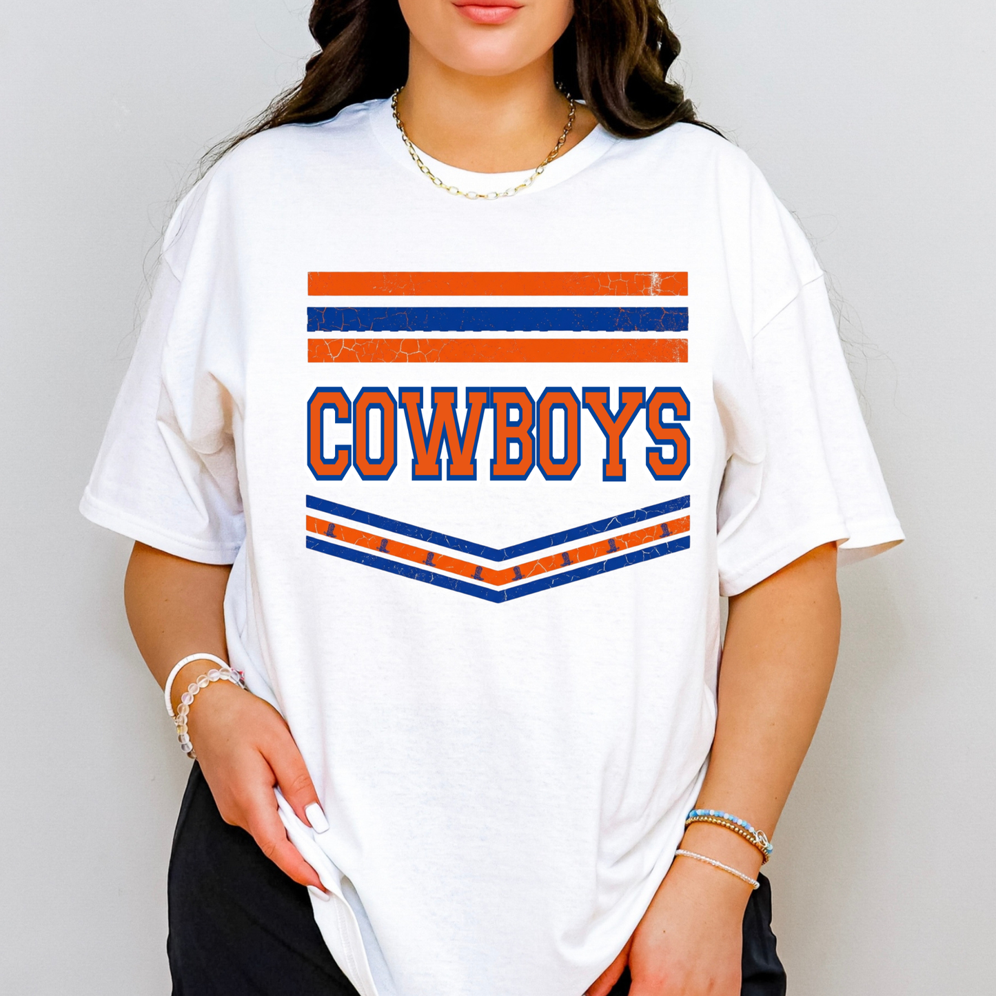 A woman wearing a white oversized graphic T-shirt with orange and blue stripes and the word 'COWBOYS' across the chest, photographed against a plain light gray background.