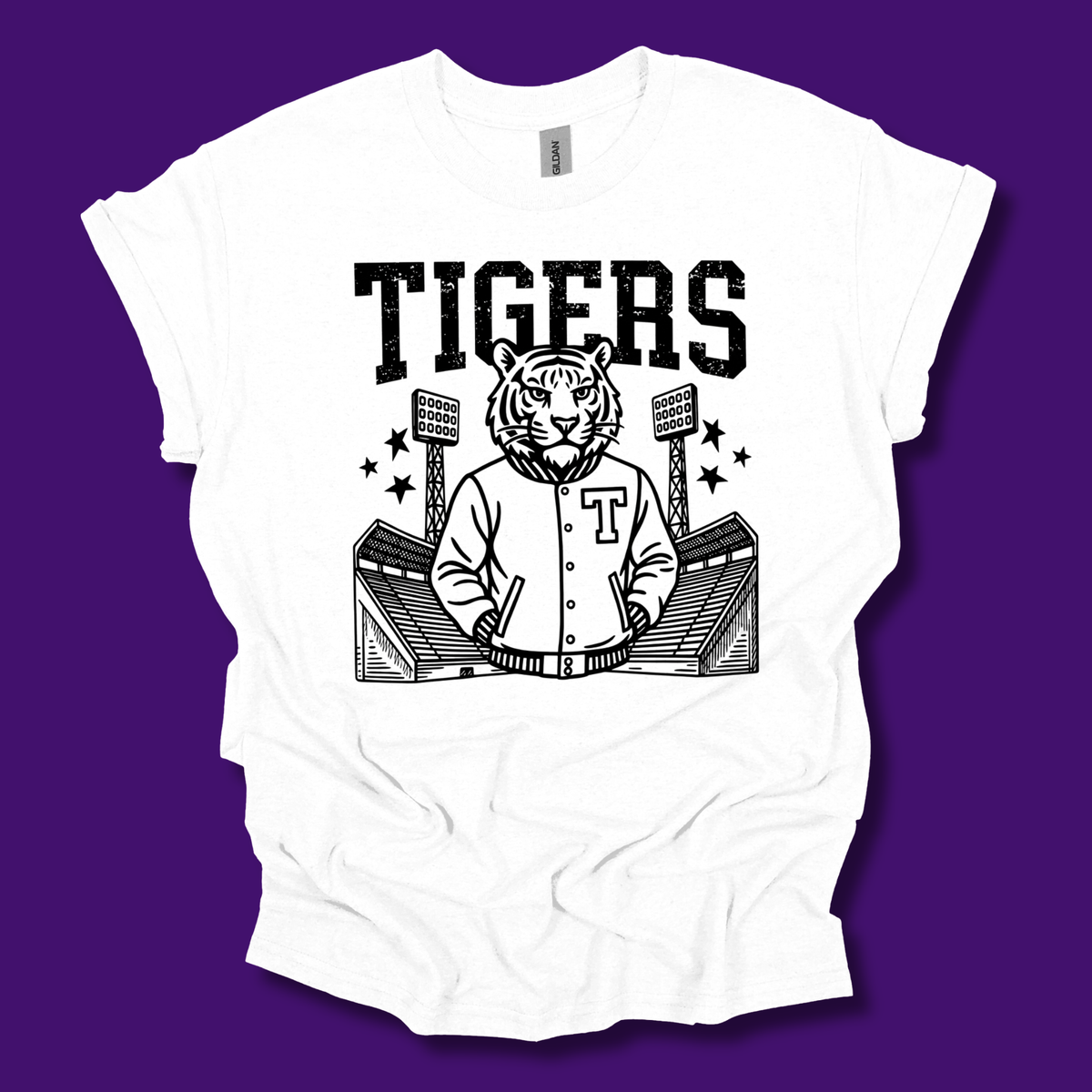 A white short-sleeve t-shirt with a black graphic print of a tiger mascot wearing a varsity jacket in front of a stadium, with the word 'TIGERS' printed above. The shirt is laid flat on a solid purple background.