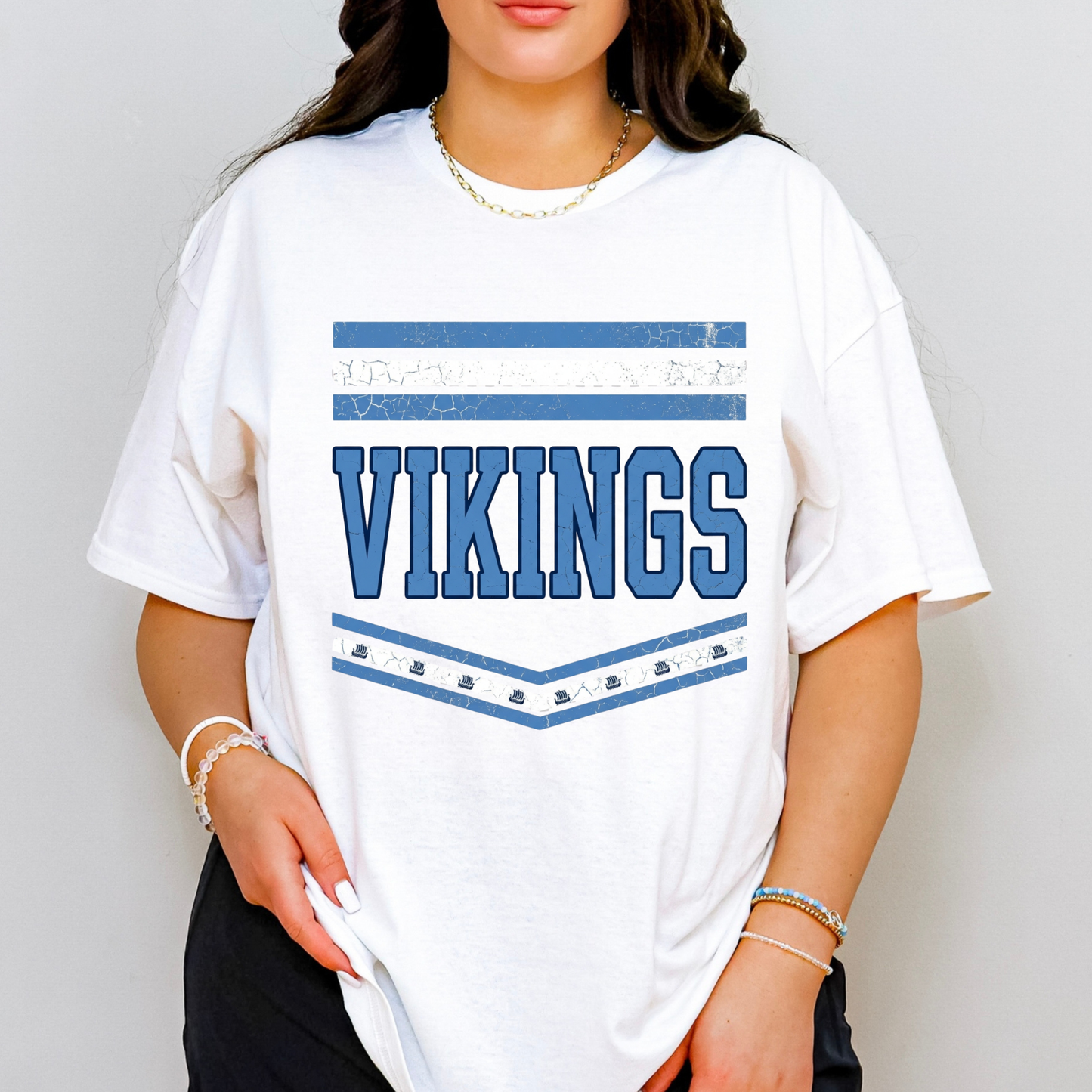 A woman wearing an oversized white t-shirt with a blue distressed graphic that reads 'VIKINGS' with horizontal stripes and small hockey pucks underneath, set against a plain light gray background.