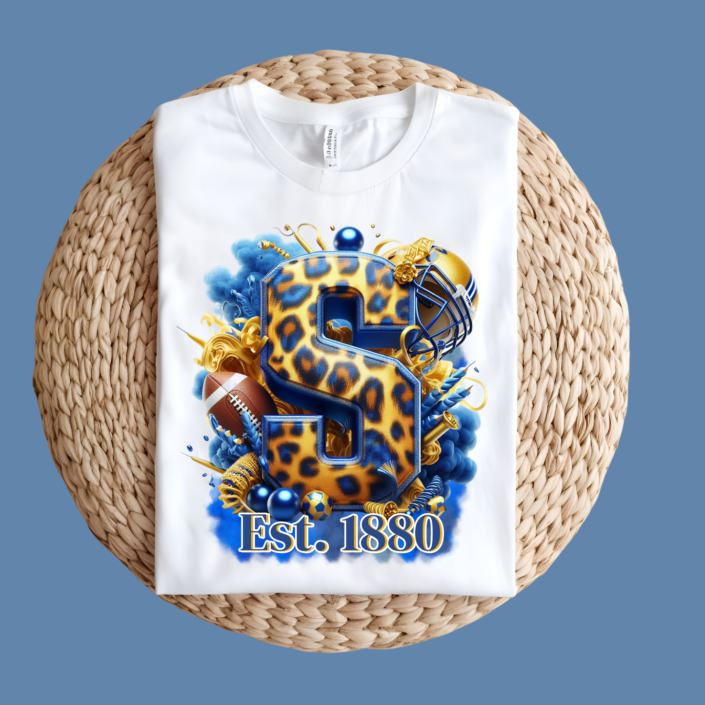 A white short-sleeve T-shirt laid flat on a round woven straw mat against a solid blue background. The shirt features a large graphic print of the number 8 in a leopard print pattern with blue outlines, surrounded by football, gold streamers, and the text 'Est.