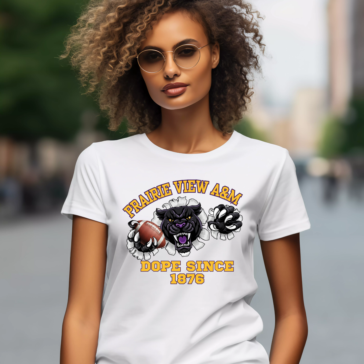 A woman wearing round gold-framed sunglasses and a white graphic t-shirt with a black panther design and the text 'PRAIRIE VIEW ARMS DOPE SINCE 1976'. The background is a blurred outdoor street scene.