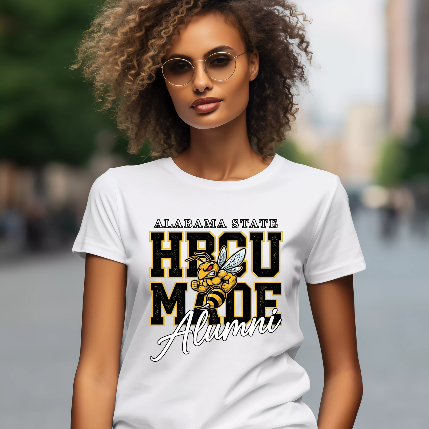A woman wearing a white graphic t-shirt with 'ALABAMA STATE HBCU MADE Alumni' and a bee illustration, set against a blurred outdoor background.