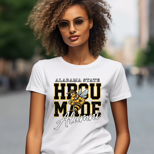 A woman wearing a white graphic t-shirt with 'ALABAMA STATE HBCU MADE Alumni' and a bee illustration, set against a blurred outdoor background.