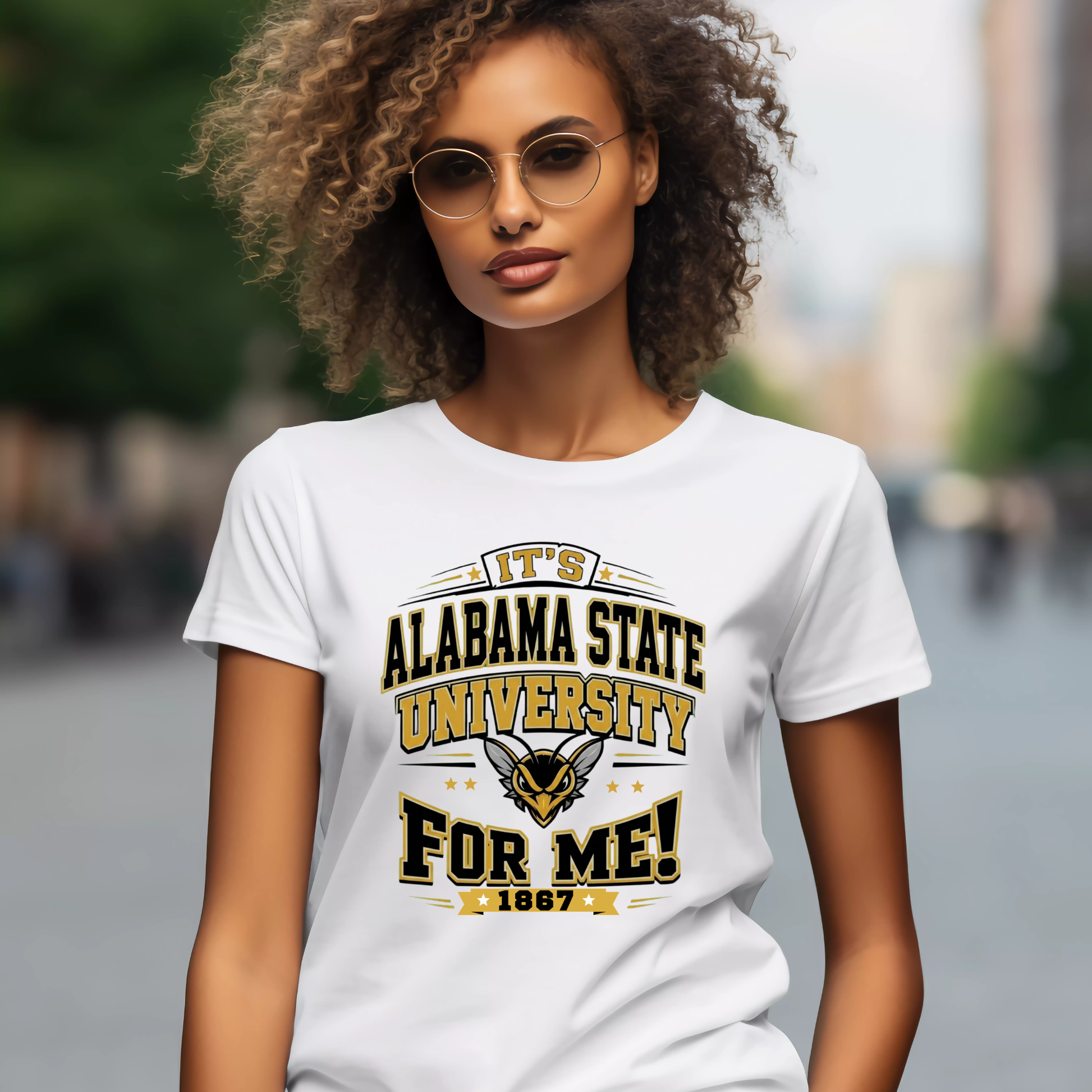 A woman with curly hair and round sunglasses wearing a white graphic t-shirt that reads 'IT'S ALABAMA STATE UNIVERSITY FOR ME! 1867' with a wildcat mascot logo, set against a blurred outdoor street background.