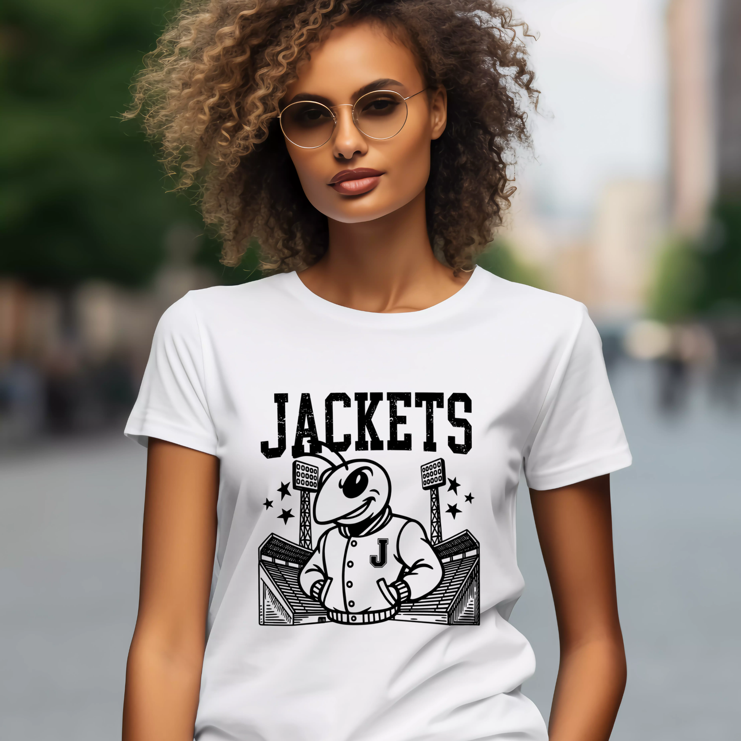 A woman wearing round sunglasses and a white graphic T-shirt with a black cartoon mascot and the word 'JACKETS' on the front, photographed outdoors with a blurred city background.