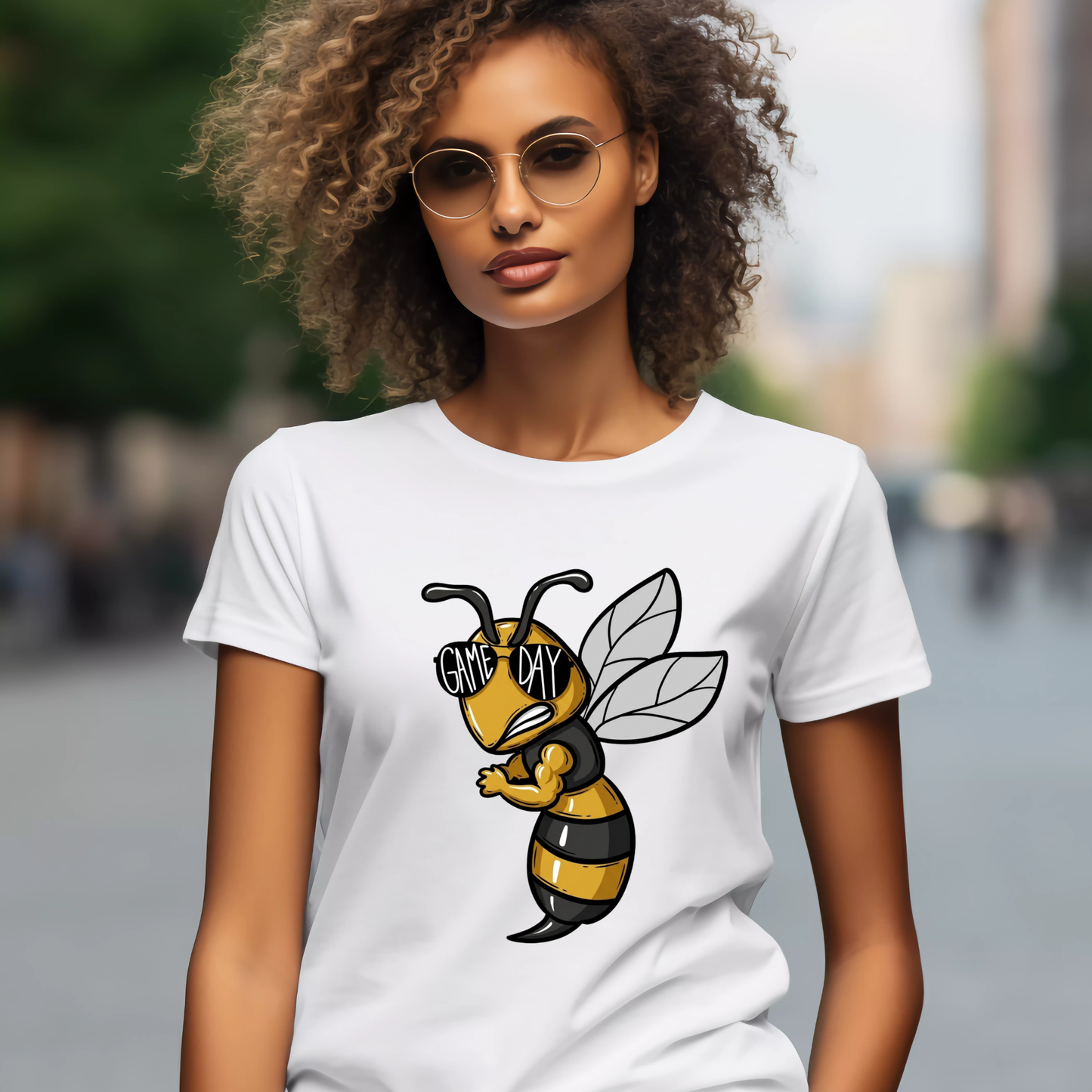 A woman wearing round sunglasses and a white graphic T-shirt with a cartoon bee design on the front, photographed outdoors with a blurred city street background.