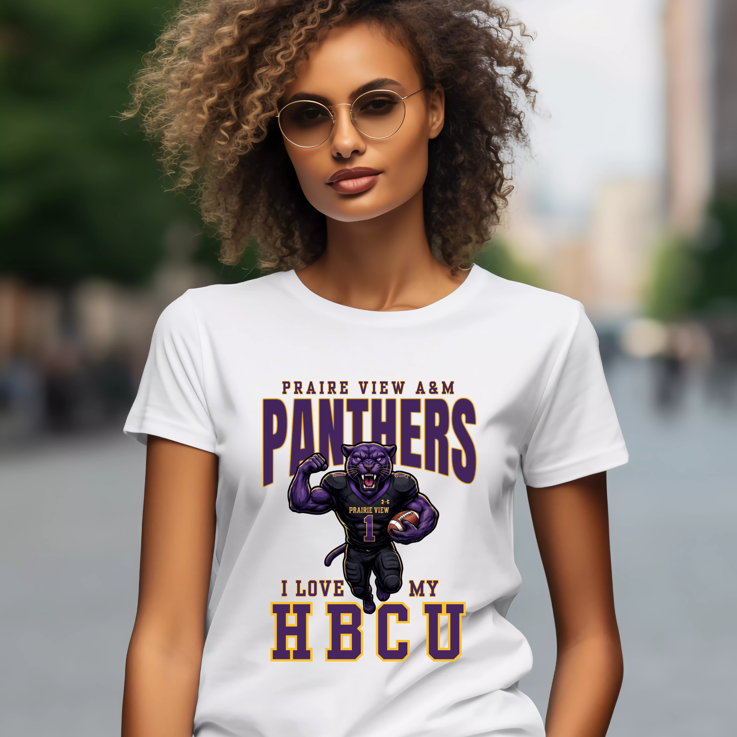 A woman wearing round glasses and a white graphic T-shirt with purple and gold text reading 'PRAIRE VIEW A&M PANTHERS I LOVE MY HBCU' and a panther mascot, photographed against a blurred outdoor street background.